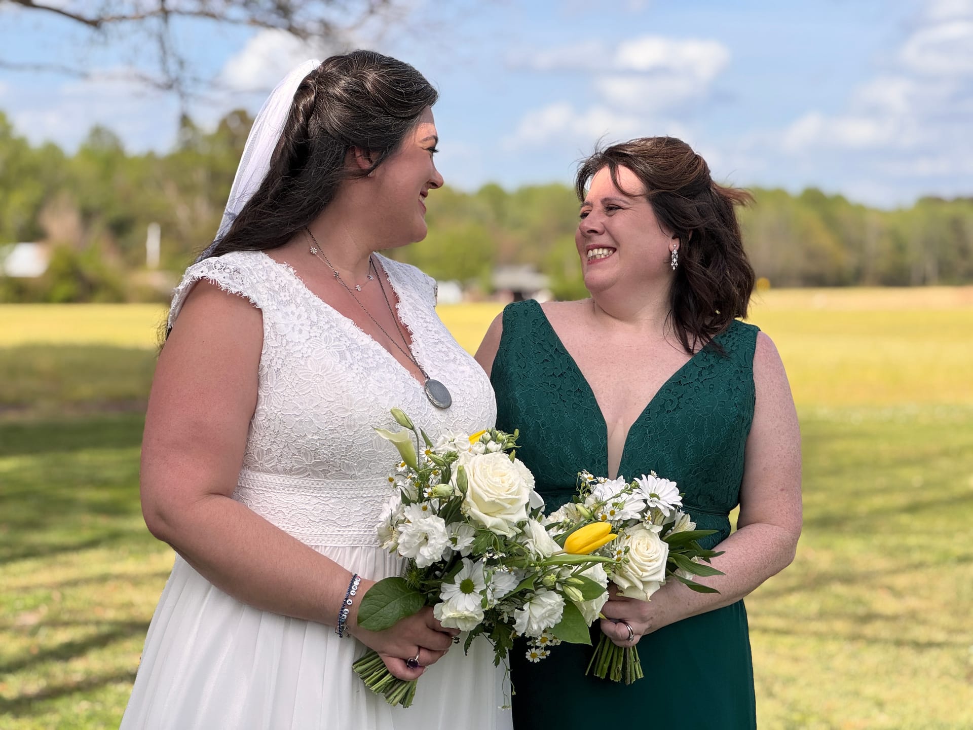 Bride in white lace gown and veil shares a joyful smile with her bridesmaid in an emerald green dress, both holding white and yellow floral bouquets against an open farmland field at Scarborough House.