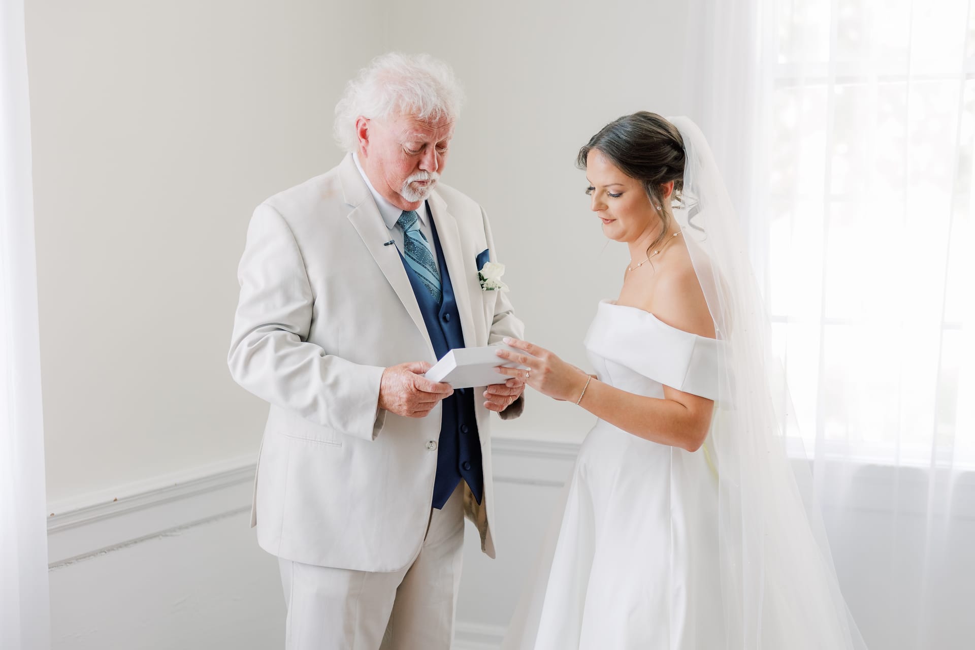 A bride in an off-shoulder white gown and veil reads a letter alongside an older gentleman in a light suit with a boutonniere inside a bright white room at Scarborough House.