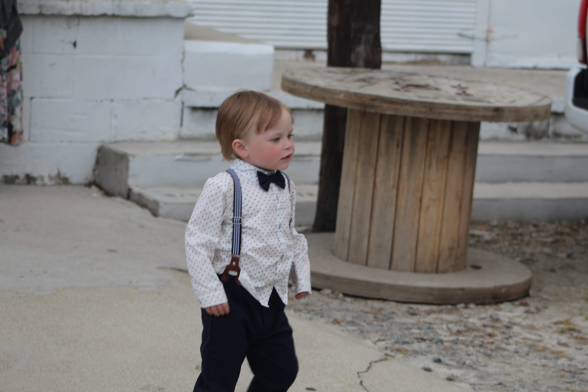 Adorable toddler ring bearer in a white patterned shirt, navy suspenders, and bow tie walks across the courtyard of a historic wedding venue.