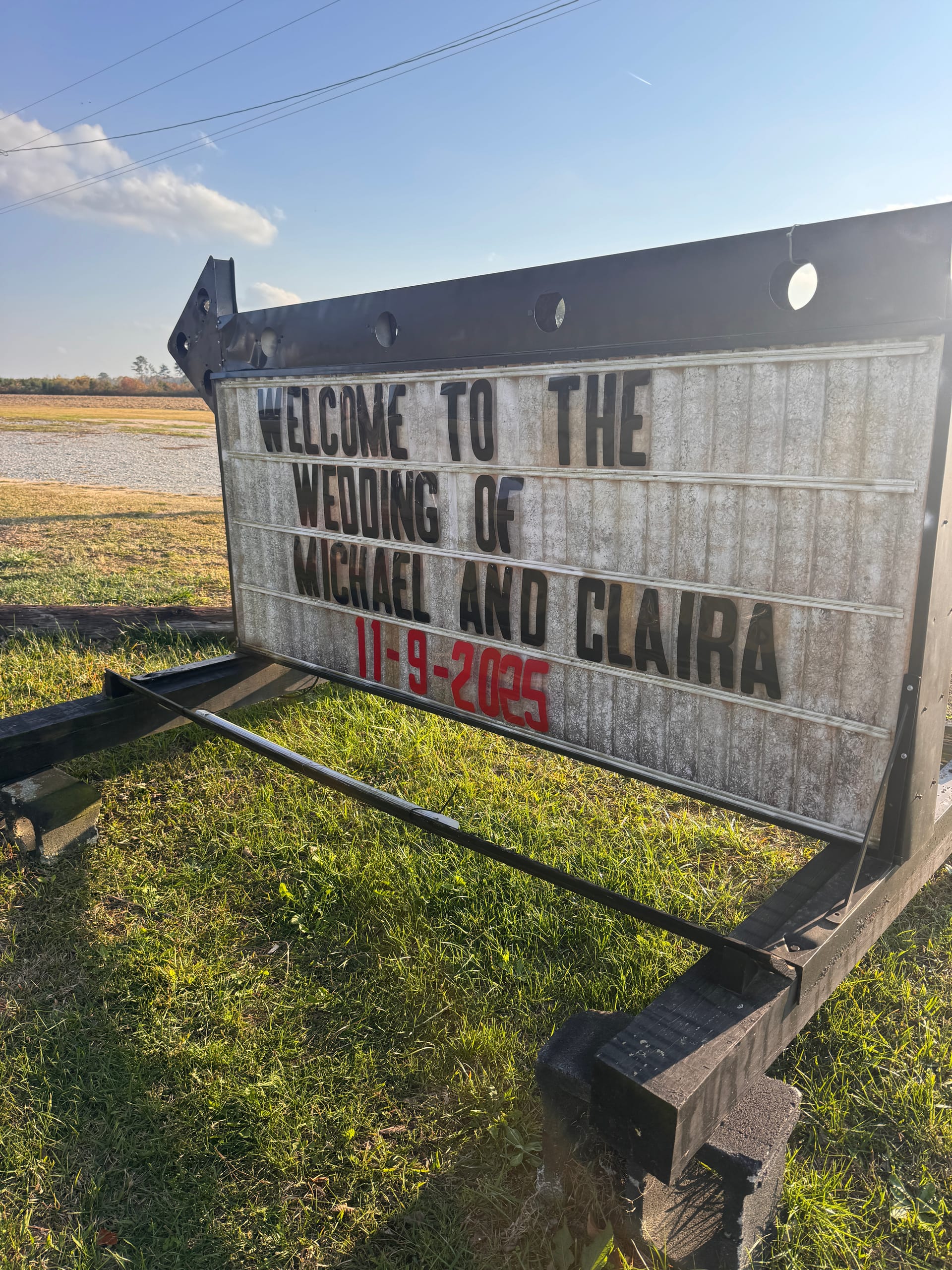Outdoor marquee sign at Scarborough House reading Welcome to the Wedding of Michael and Claira 11-9-2025 set against open farmland fields and blue sky.