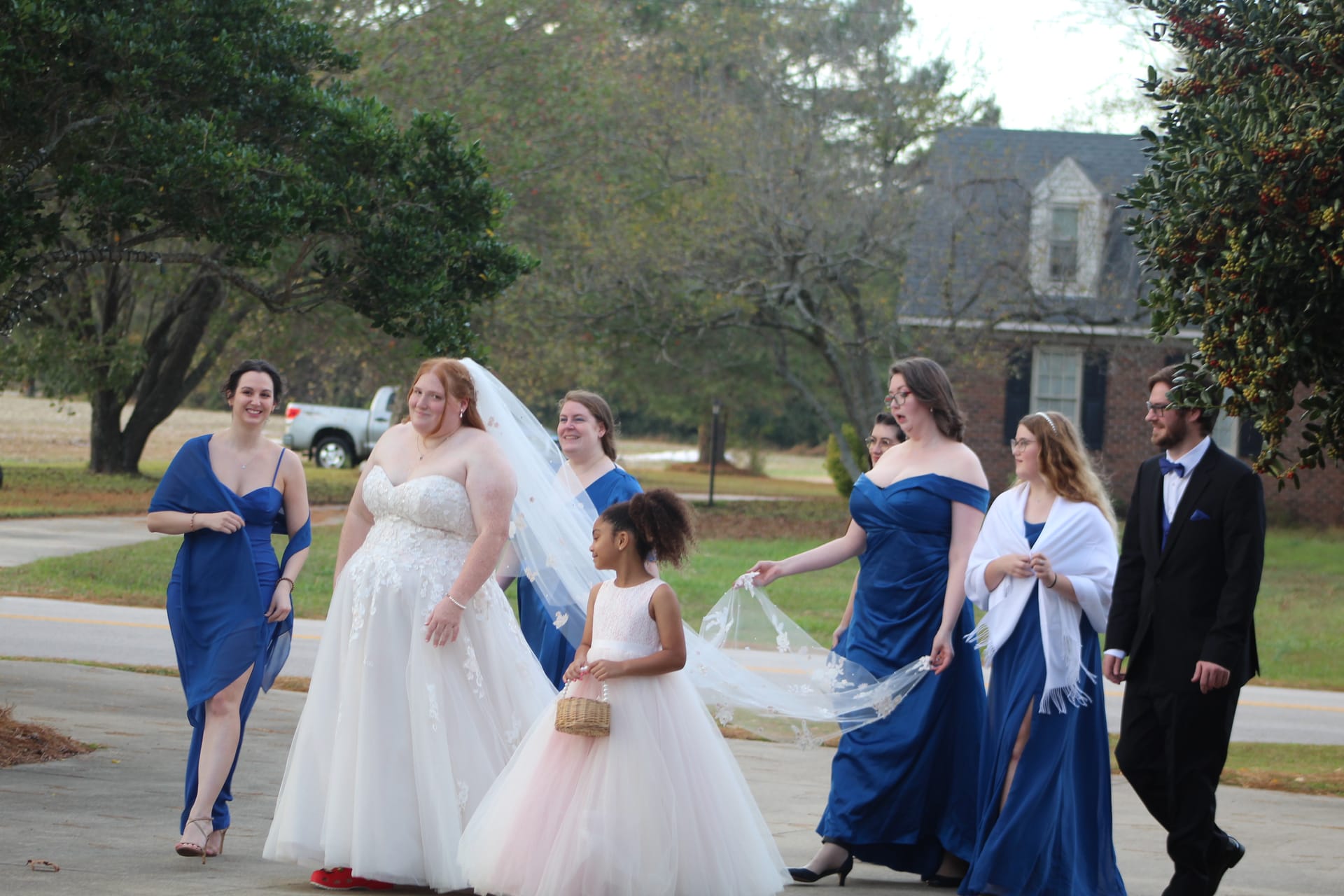 Bride in a lace ballgown walks alongside her bridesmaids in royal-blue gowns and a young flower girl carrying a basket, with a historic manor house visible in the background at Scarborough House.