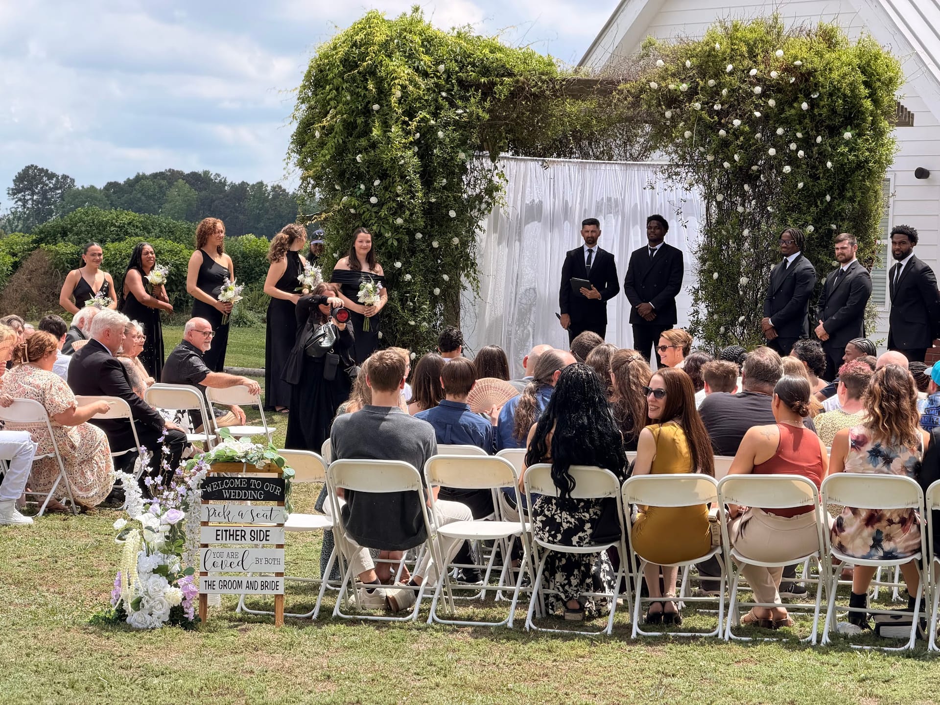 Outdoor wedding ceremony on the Scarborough House lawn with guests seated in white folding chairs facing an ivy-draped arch adorned with white florals, bridesmaids in black dresses and groomsmen in suits standing at the altar.