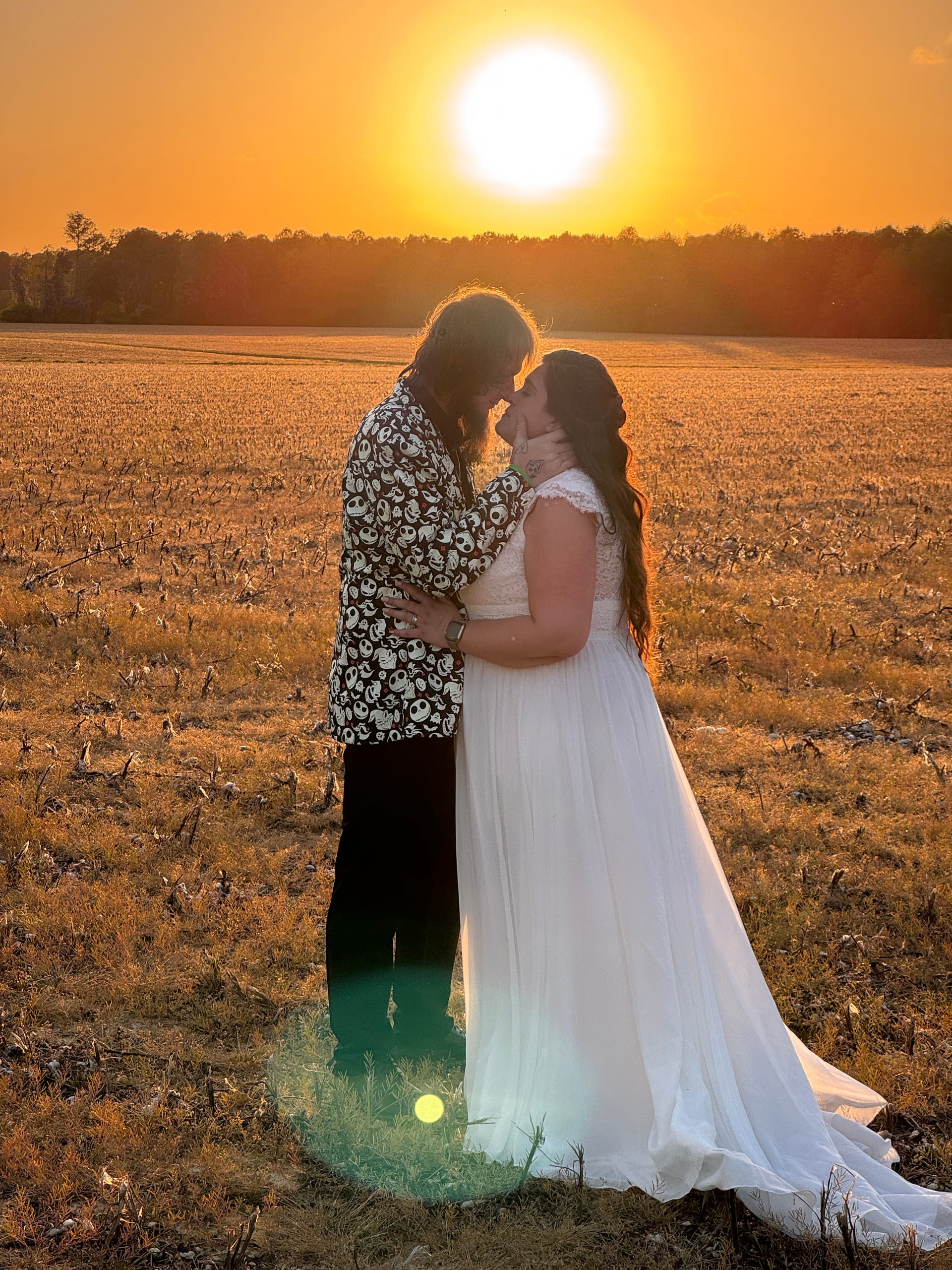 Bride in a flowing white gown and groom in a patterned blazer share an intimate almost-kiss silhouetted against a vibrant golden sunset over open farmland fields at Scarborough House.