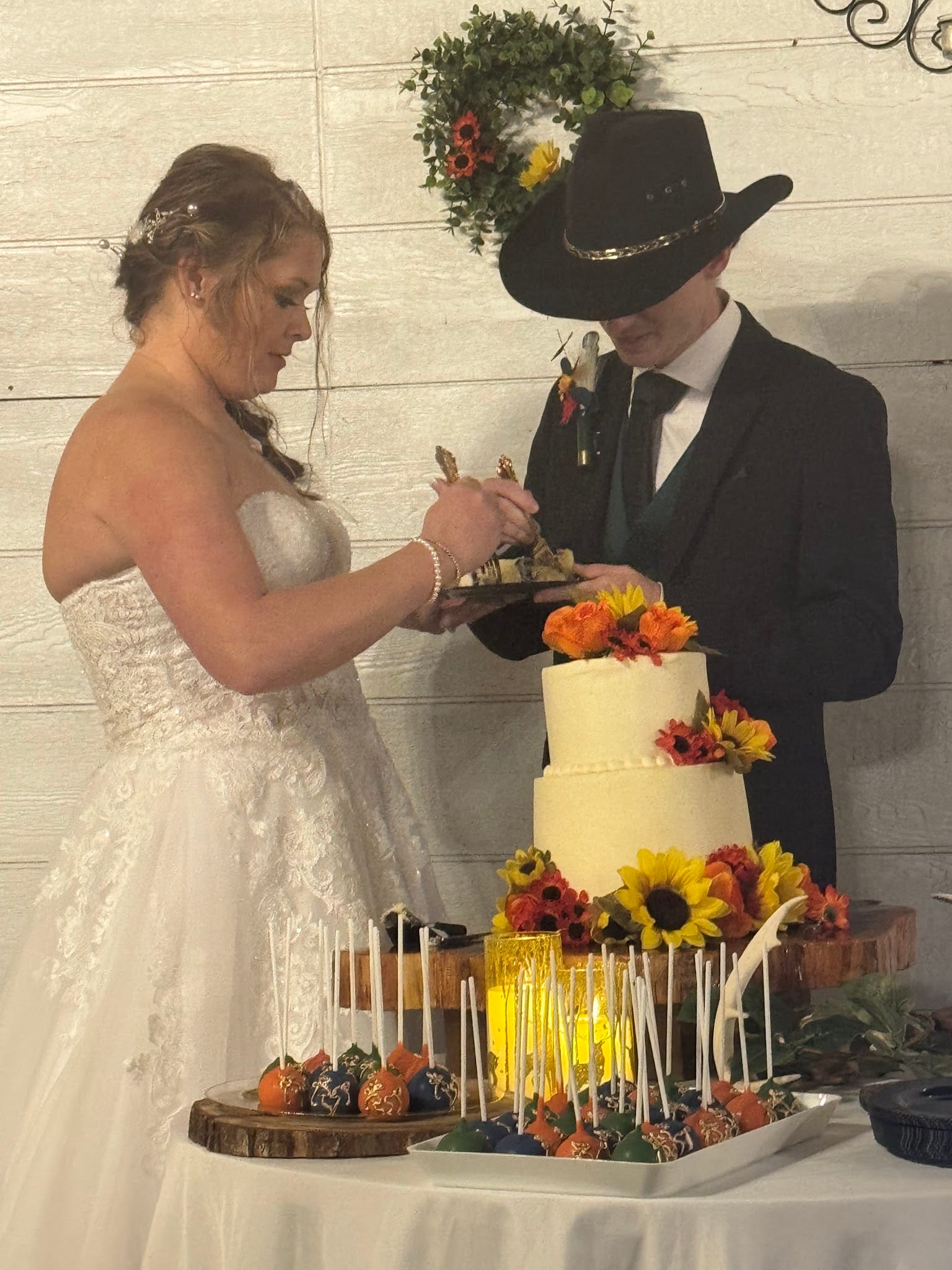 Bride in a white lace gown and groom in a dark suit and cowboy hat cutting their two-tier wedding cake adorned with sunflowers and autumn florals at a rustic estate reception.