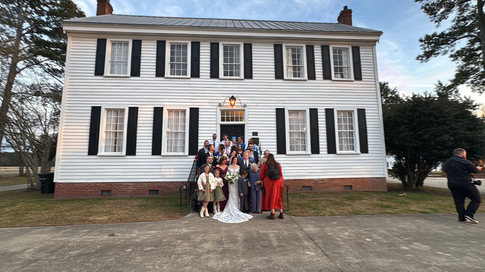 Wedding party and family gathered on the front steps of the white clapboard 1821 Scarborough House manor with black shutters at dusk.
