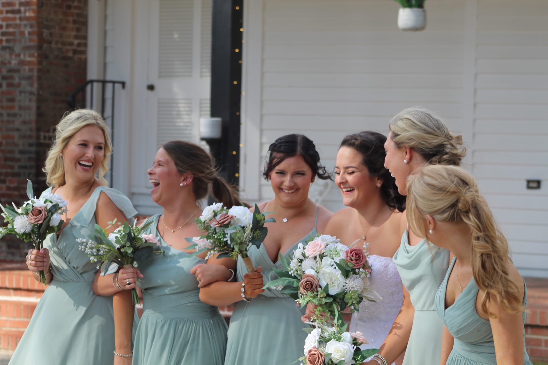 Bride in a lace gown laughs with five bridesmaids in sage green dresses holding blush and white bouquets outside Scarborough House historic manor.
