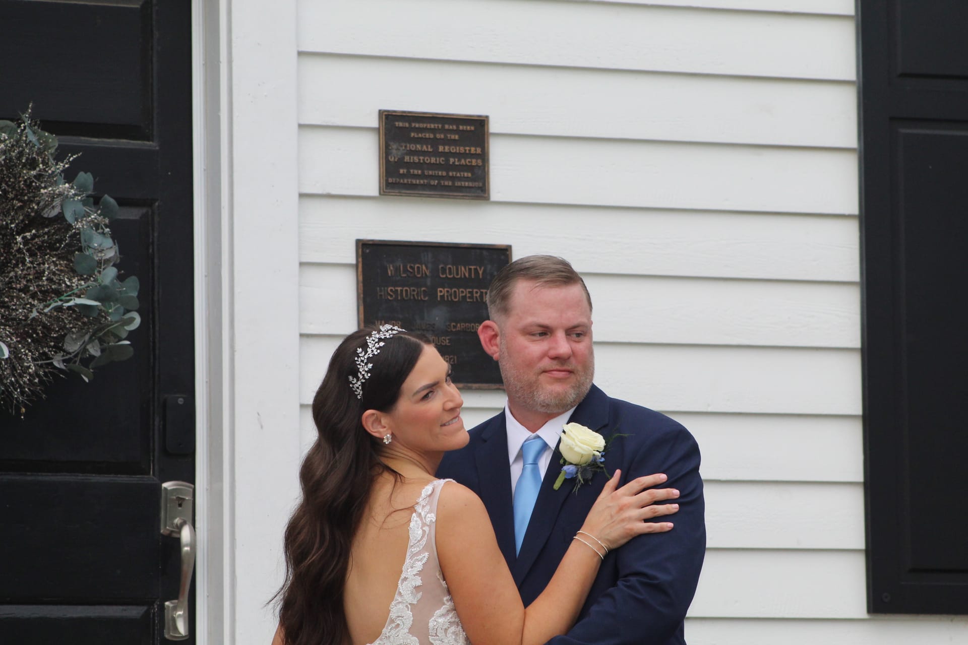 Bride in lace gown with floral hairpiece gazes at groom in navy suit and light blue tie outside the 1821 Wilson County Historic Property manor house at Scarborough House.