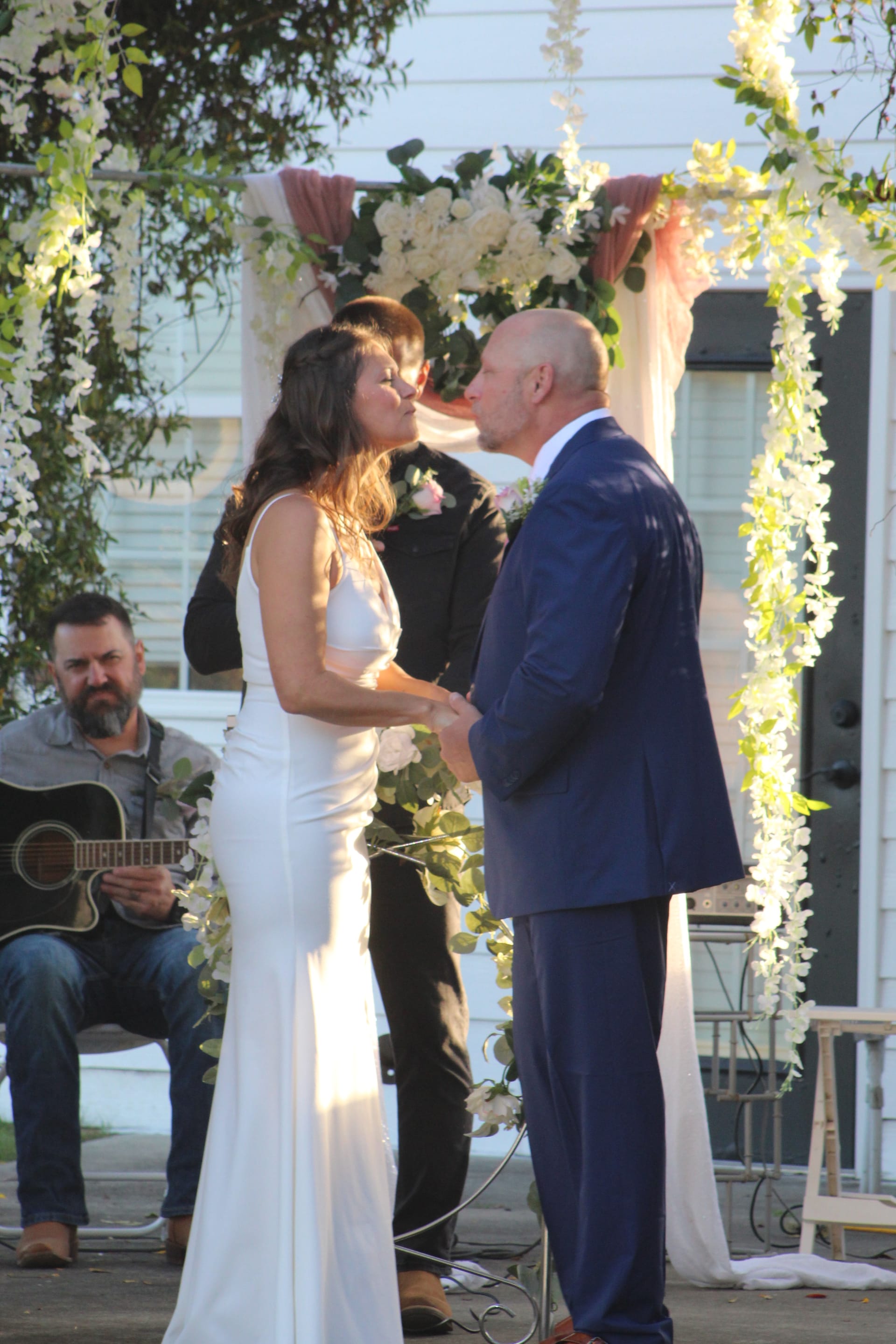 Bride and groom share their first kiss beneath a lush floral arch adorned with white wisteria and pink roses while a guitarist plays in the background at Scarborough House.