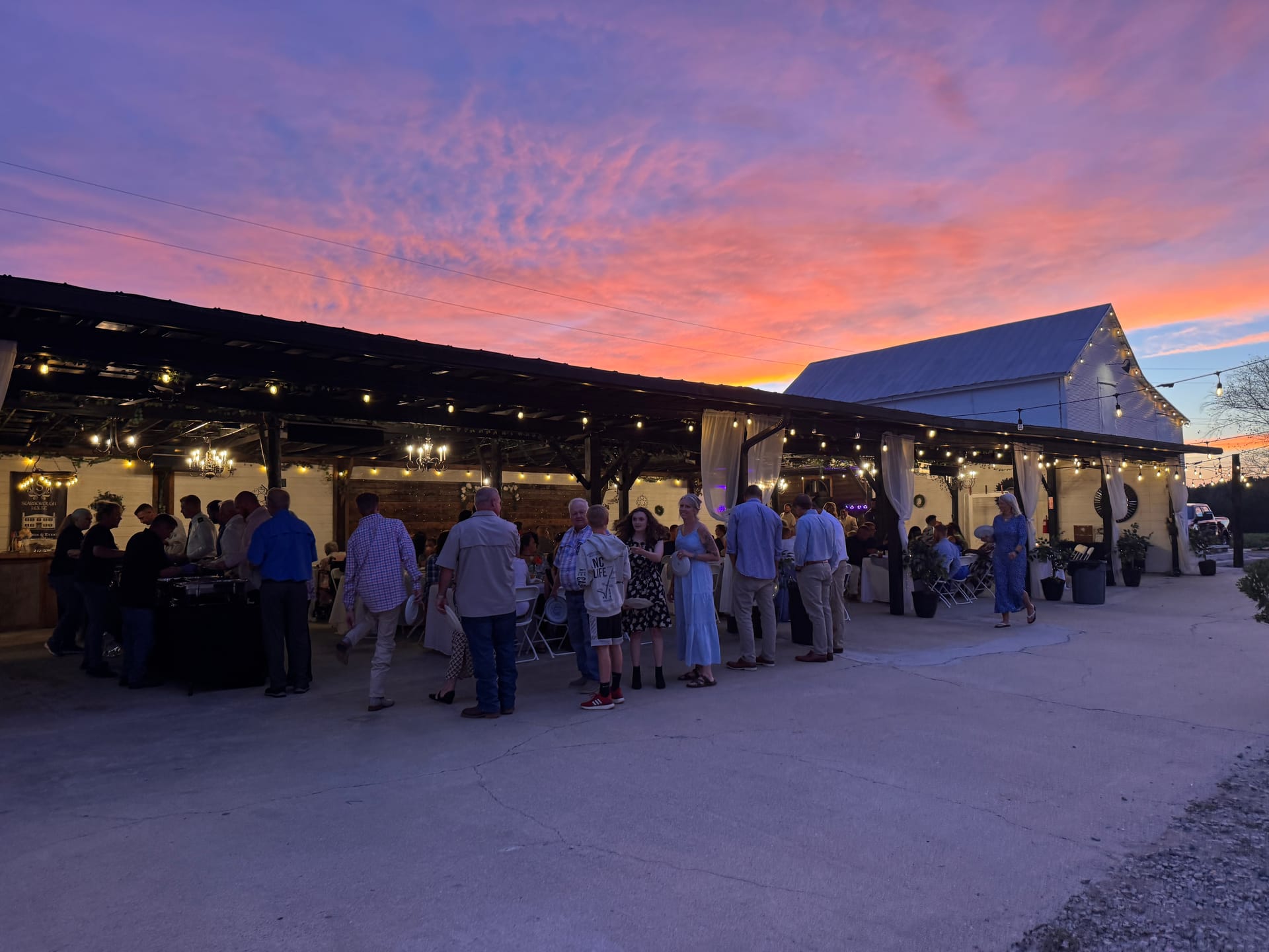 Guests mingle under a string-lit open-air pavilion at Scarborough House as a dramatic pink and orange sunset fills the sky behind the white barn venue.