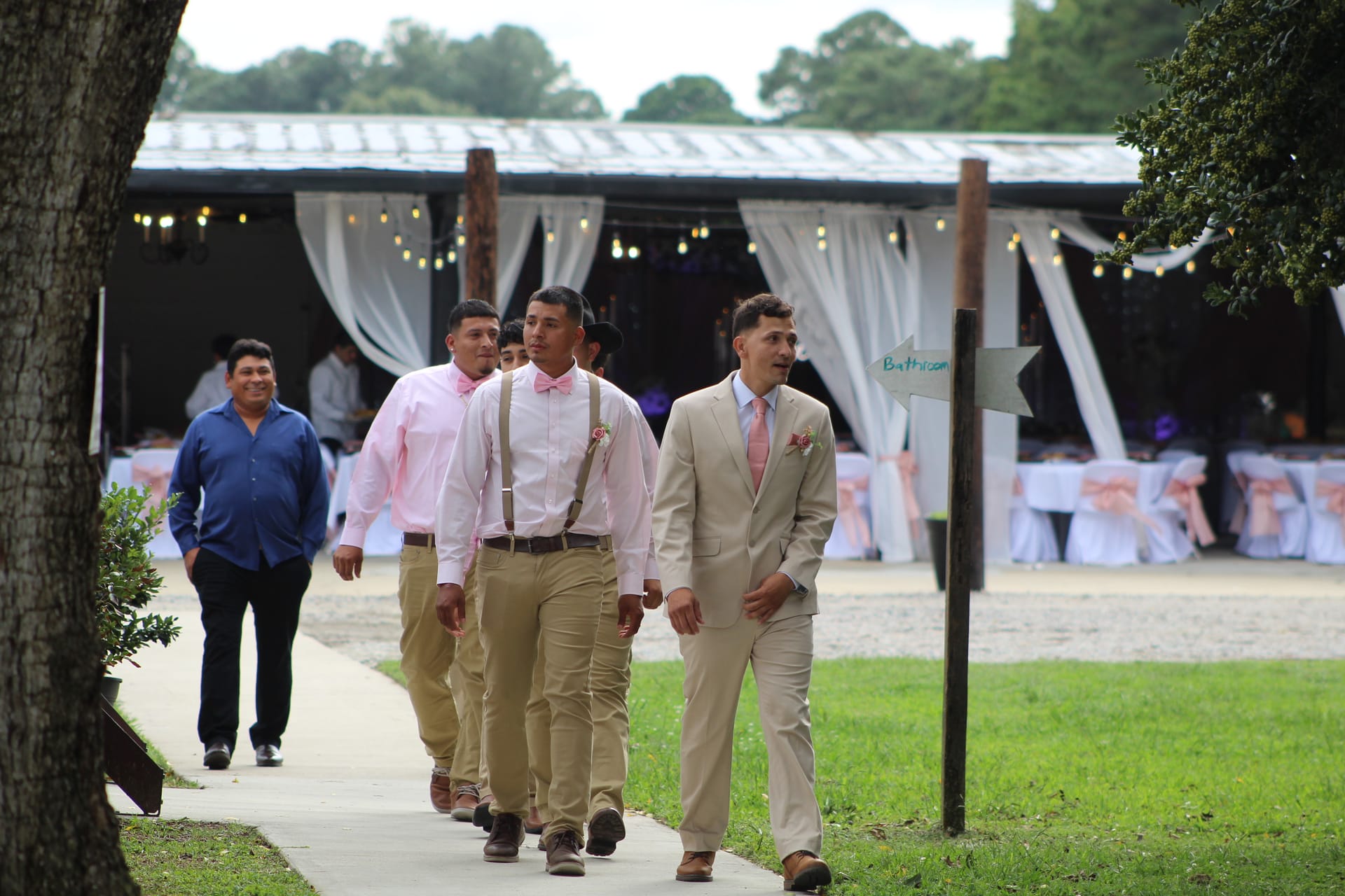 Groom in a tan suit leads his groomsmen in pink shirts and suspenders along a pathway toward the decorated outdoor pavilion at Scarborough House wedding venue.