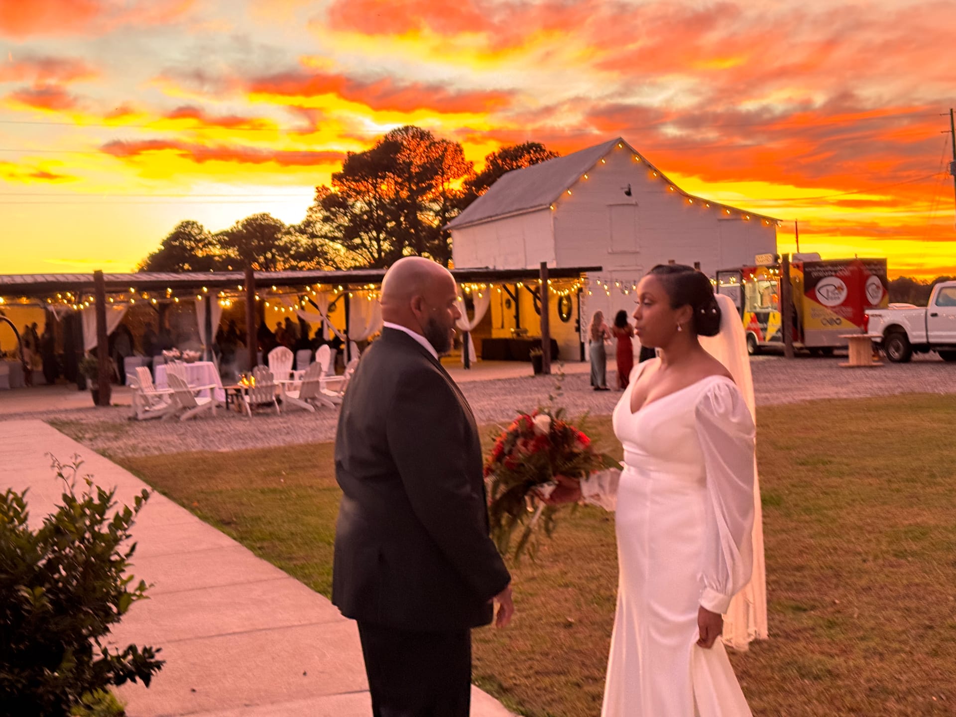 Bride in an elegant white off-shoulder gown holding a red bouquet faces her groom in a dark suit against a dramatic orange and red sunset sky with the Scarborough House white barn and string lights glowing in the background.