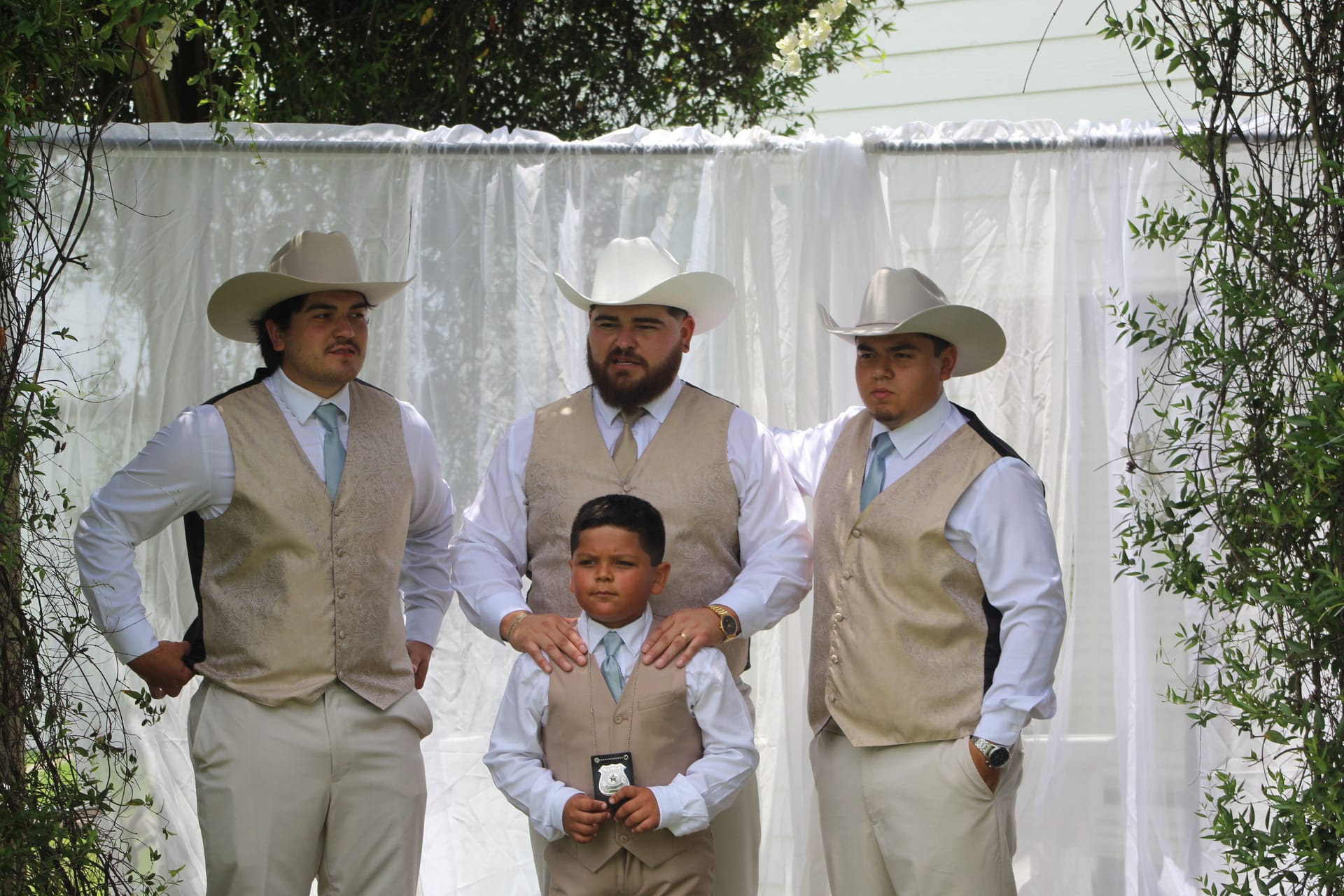 Groom and two groomsmen wearing tan vests and white cowboy hats stand before a draped ceremony backdrop with a young ring bearer holding a badge at an outdoor western-style wedding.