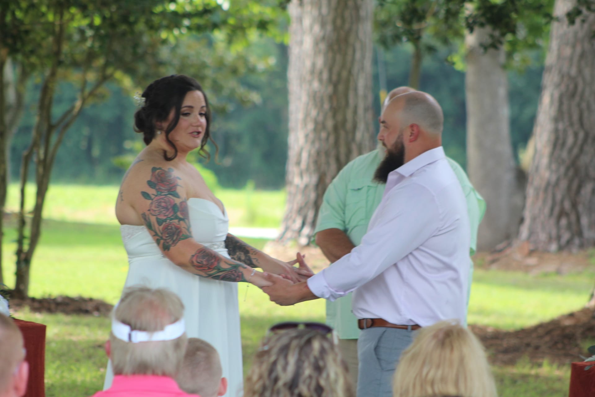 Tattooed bride in a strapless white gown holds hands with her bearded groom during outdoor wedding vows beneath towering oak trees at Scarborough House estate.