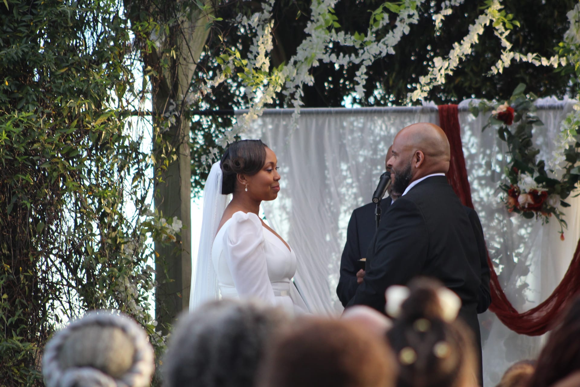 Bride in a white gown and veil exchanges vows with her groom beneath a floral arch adorned with white blooms and burgundy draping at an outdoor garden ceremony.