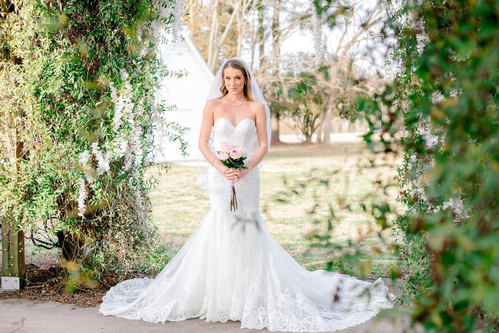 Bride in a strapless lace mermaid wedding gown and veil holding a pink rose bouquet, framed by a lush floral and greenery archway at Scarborough House historic estate.