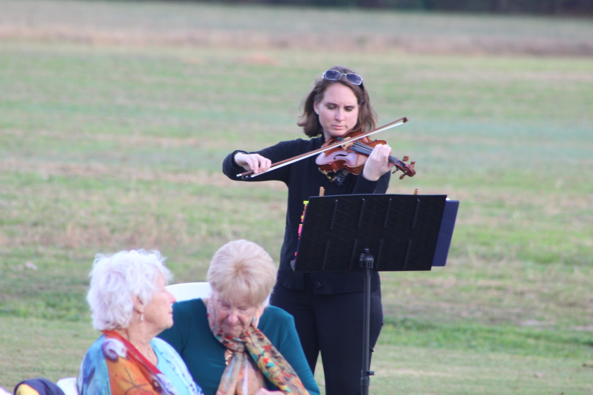 A violinist in black plays from sheet music on a stand during an outdoor wedding ceremony on the lawn at Scarborough House while two seated elderly guests listen.