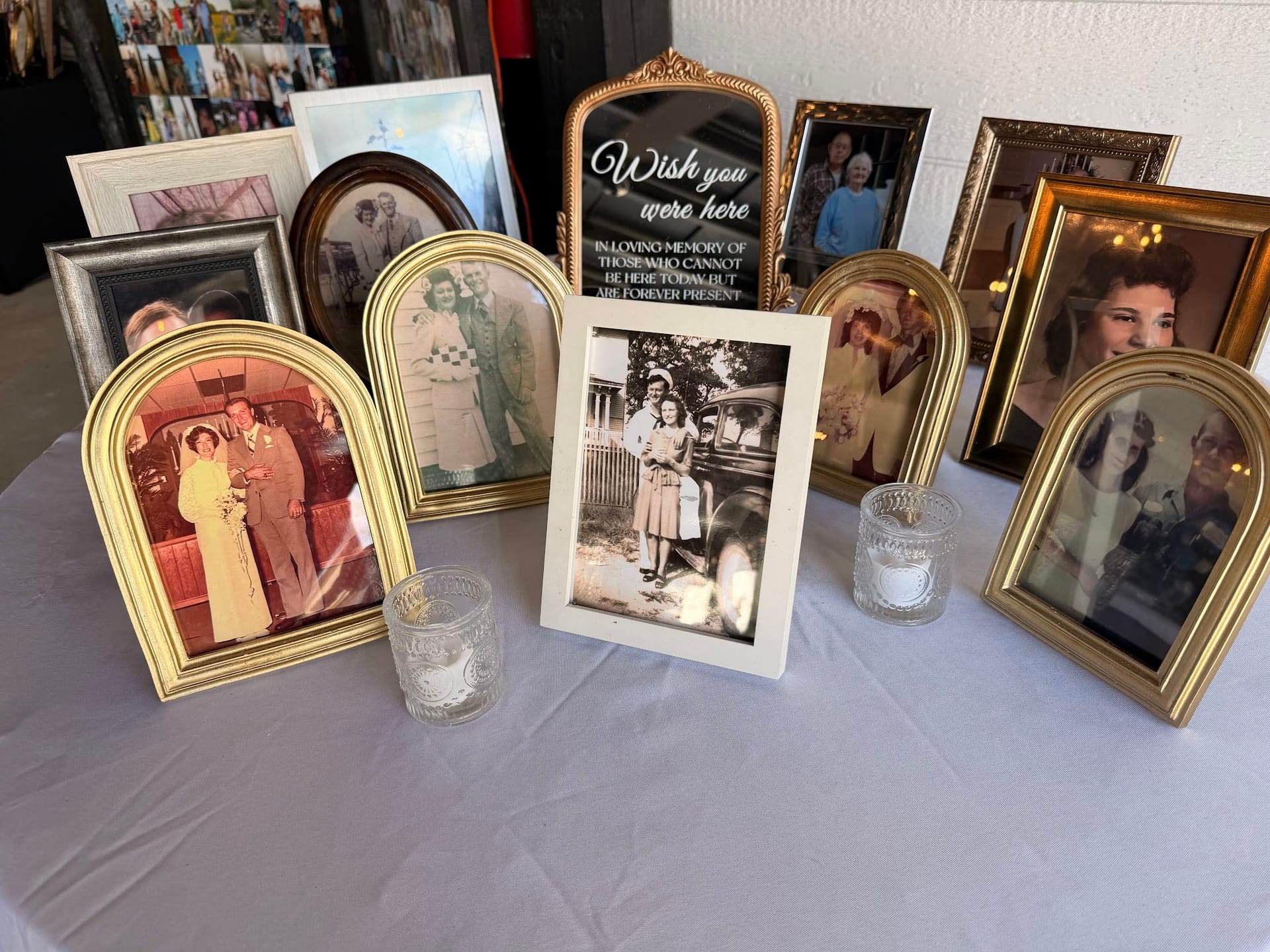 A memorial table display at a wedding featuring gold-framed vintage photographs of loved ones, small crystal votive candles, and a decorative sign reading Wish You Were Here in loving memory of those who cannot be here today.