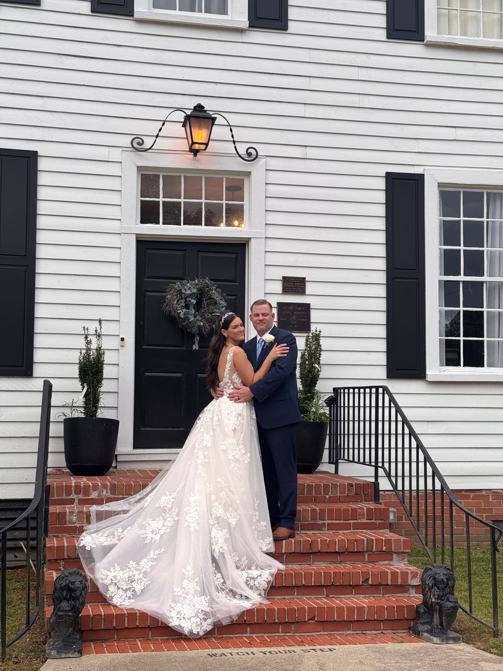 Bride in a flowing lace ball gown and groom in a navy suit embrace on the red brick front steps of the 1821 Scarborough House manor, framed by a black door with eucalyptus wreath and a glowing iron lantern.