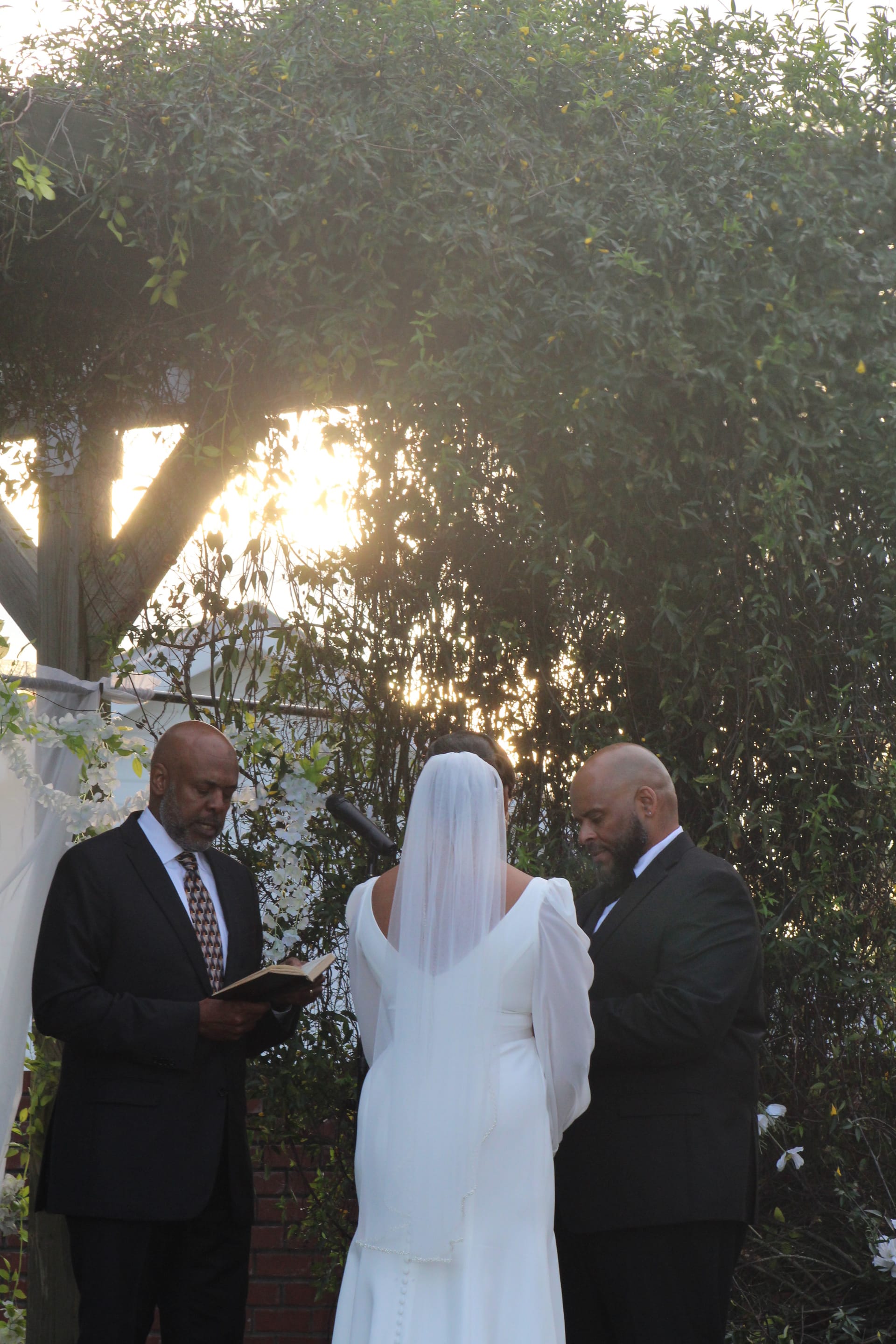 A bride in a white gown and veil stands with her groom and officiant during an outdoor garden ceremony bathed in warm golden-hour light beneath a canopy of lush trees.