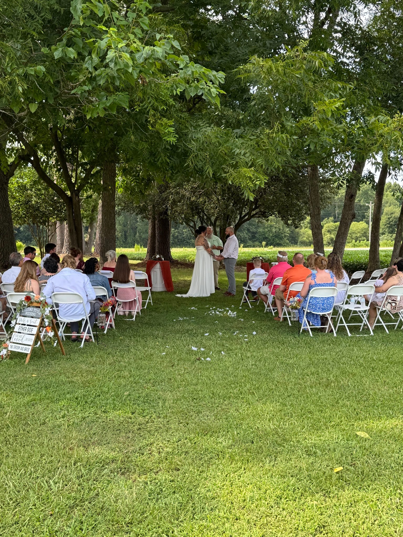 Guests seated in white folding chairs on a shaded lawn watch a bride and groom exchange vows beneath tall trees at a historic outdoor wedding ceremony venue.
