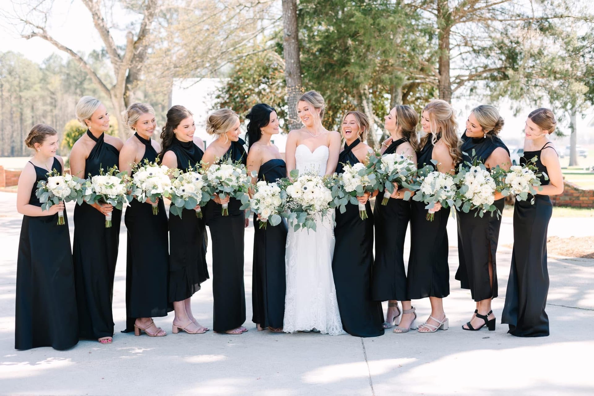 Bride in a white lace gown laughing joyfully with eleven bridesmaids in black gowns holding white floral bouquets outdoors at Scarborough House wedding venue.