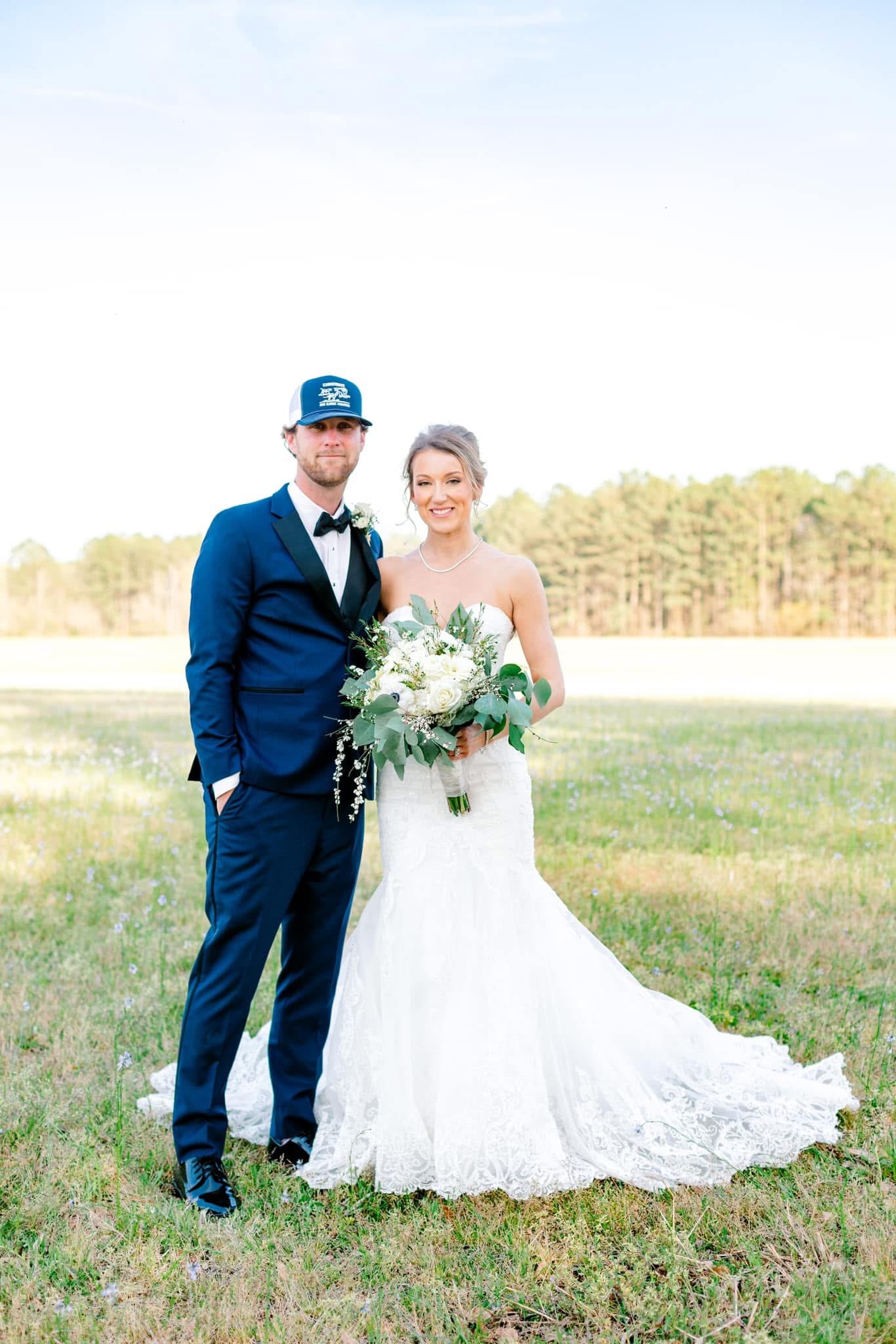 Bride in a lace mermaid gown holding a white and greenery bouquet stands with her groom in a navy tuxedo and blue trucker cap in an open field at Scarborough House estate.