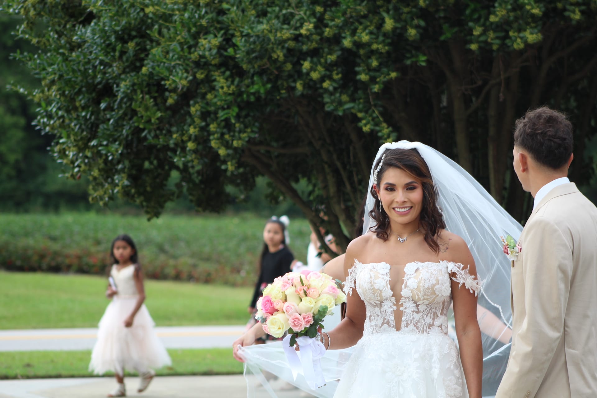 Smiling bride in off-shoulder lace gown holding pink and ivory rose bouquet walks toward groom beneath towering oak trees at Scarborough House estate wedding venue.