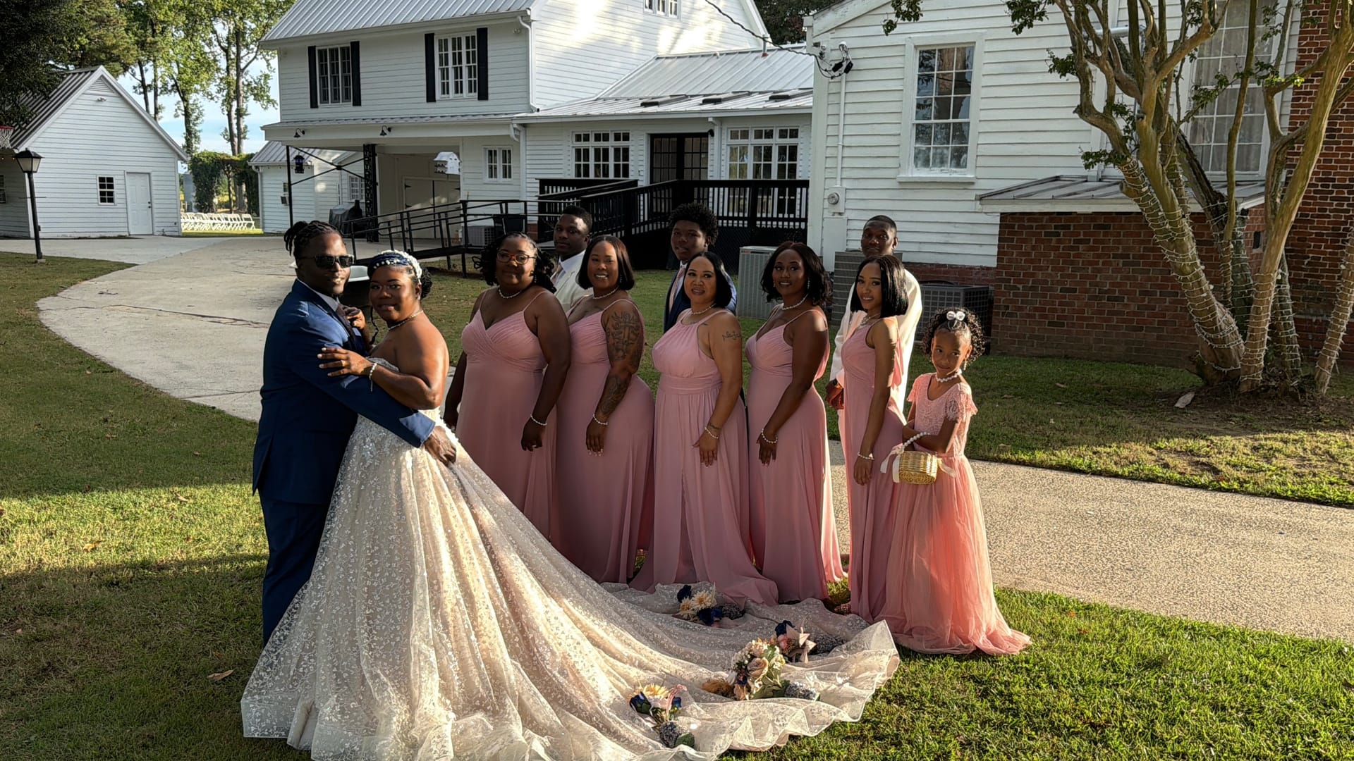 Bride in an elaborate white ballgown with a sweeping train poses with her groom in a navy suit and bridesmaids in blush pink dresses in front of the historic white manor house at Scarborough House.