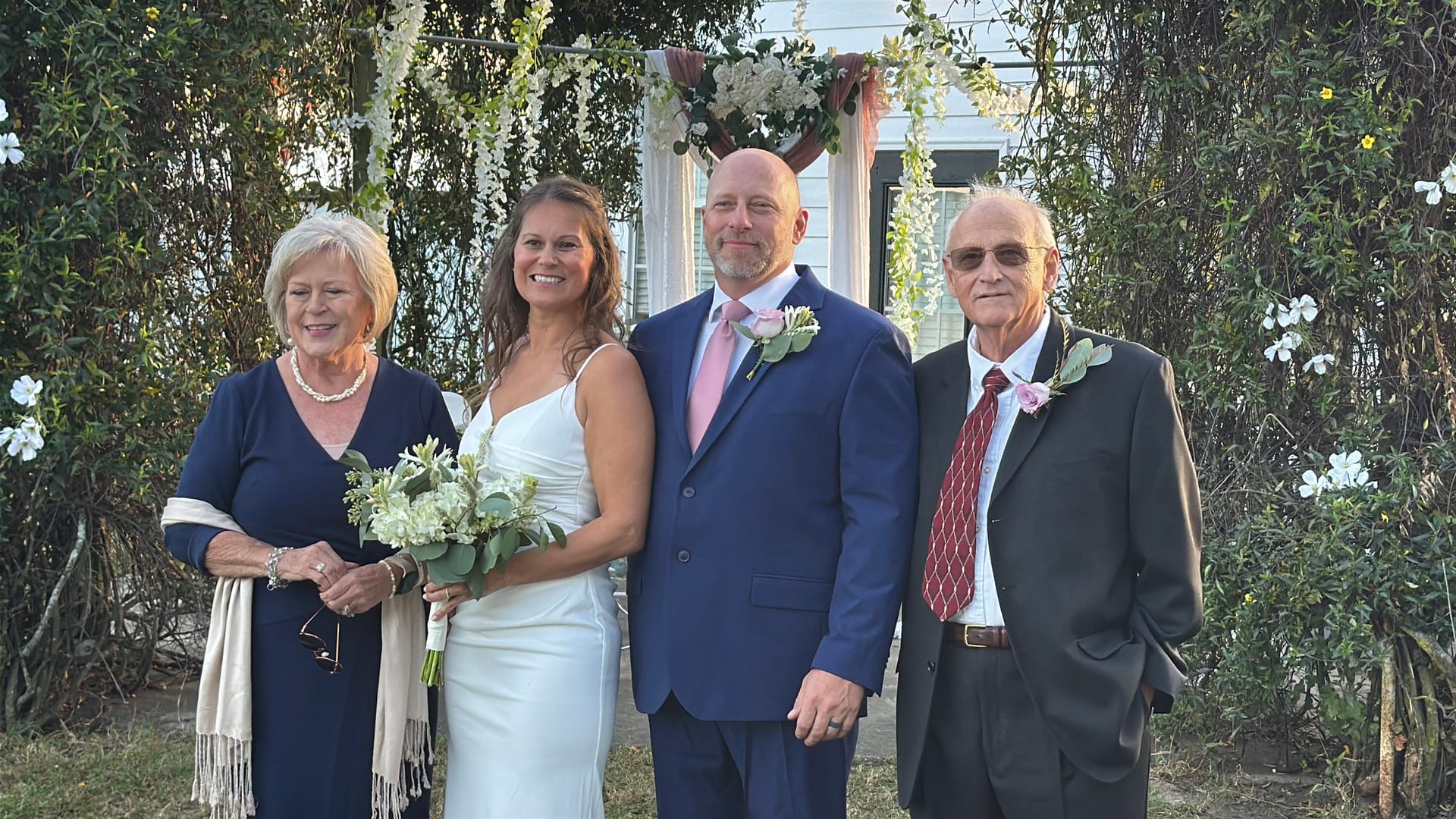 Bride in white gown holding a white bouquet stands with groom in navy suit and two family members beside a floral arch at Scarborough House wedding venue.