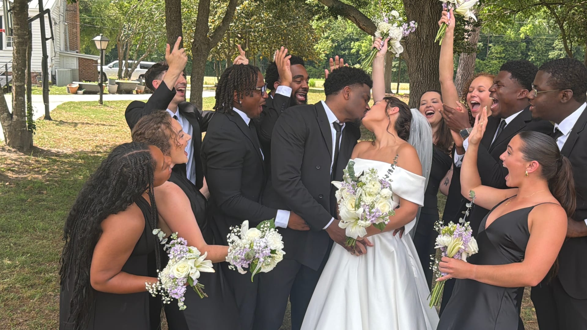 Bride and groom share a kiss while their wedding party cheers and raises bouquets beneath large oak trees at Scarborough House.