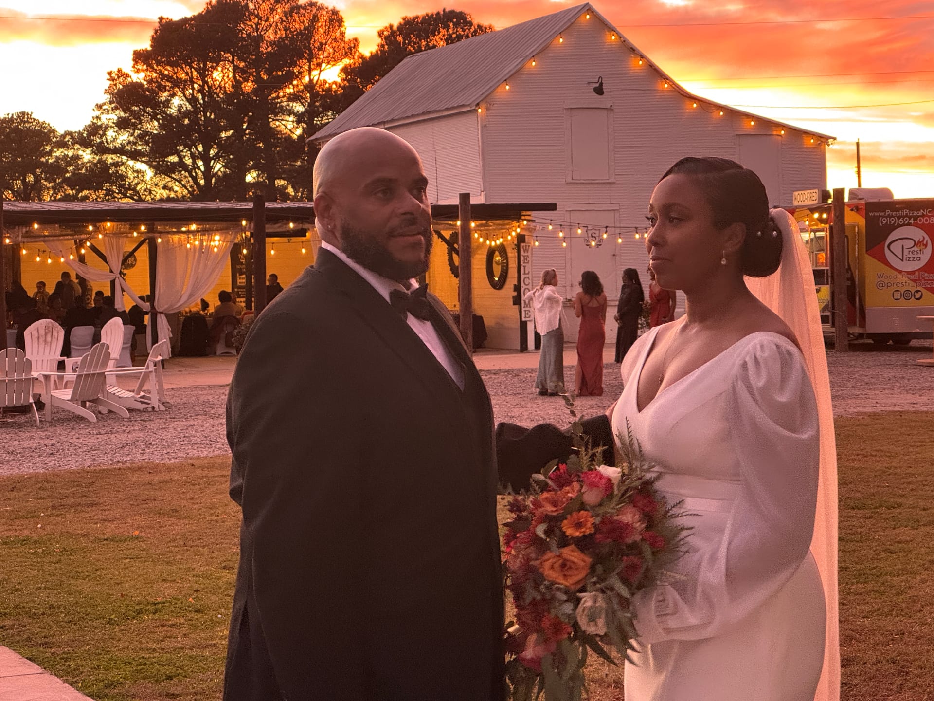 Bride in a long-sleeve white gown holding a fall floral bouquet stands beside her tuxedoed groom in front of a white barn venue under a dramatic fiery orange sunset sky.
