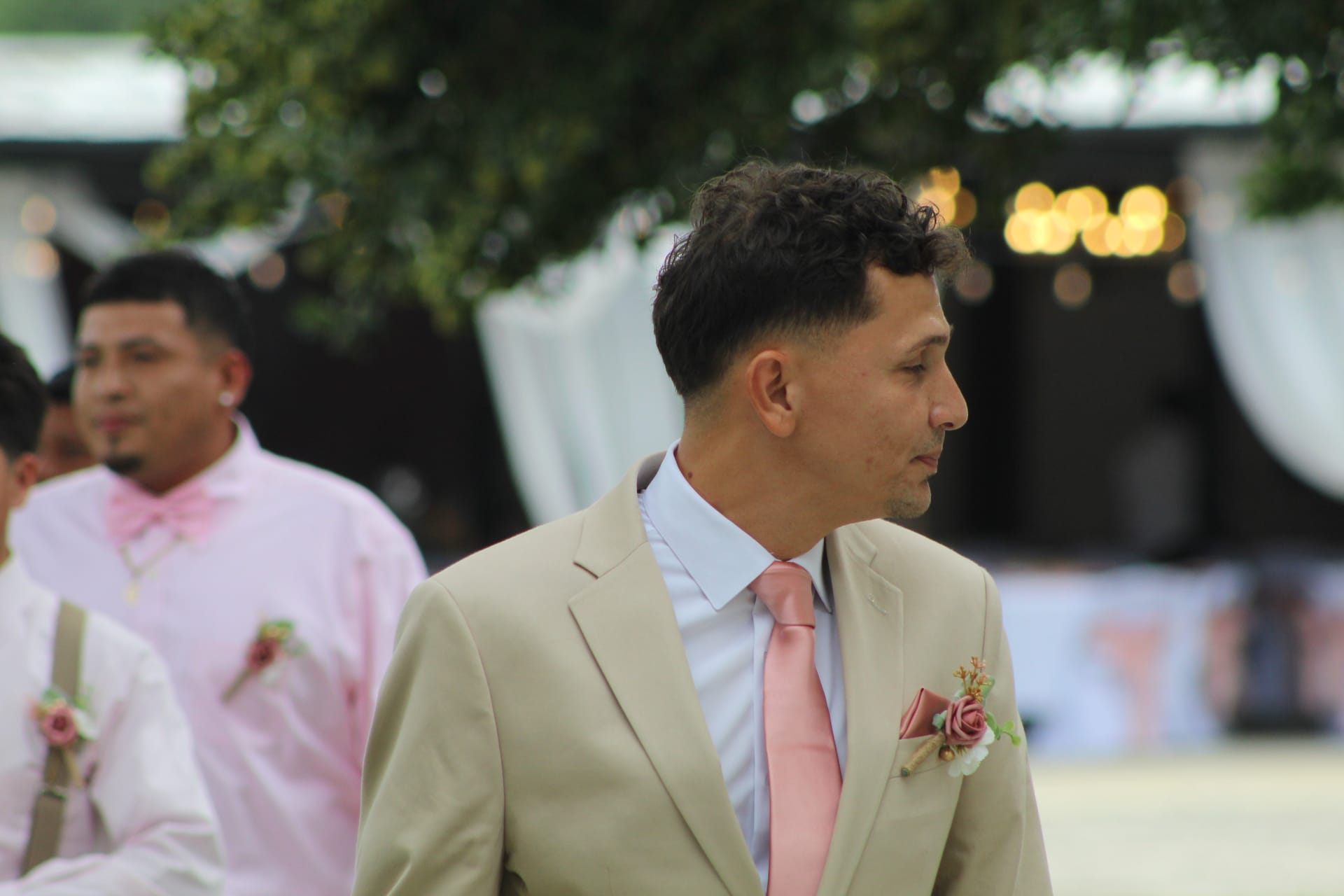 Groom in a tan suit with a pink tie and floral boutonniere stands in profile at an outdoor wedding venue with other groomsmen visible in the background.