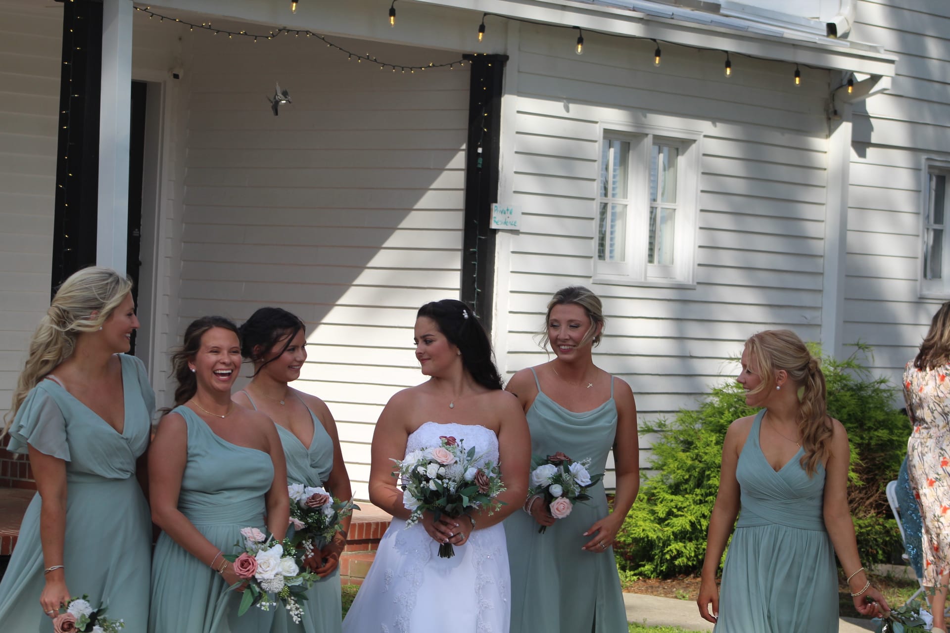 Bride in white gown holding a pink and white floral bouquet laughing and sharing a joyful moment with five bridesmaids in sage green dresses outside the historic white clapboard facade of Scarborough House.