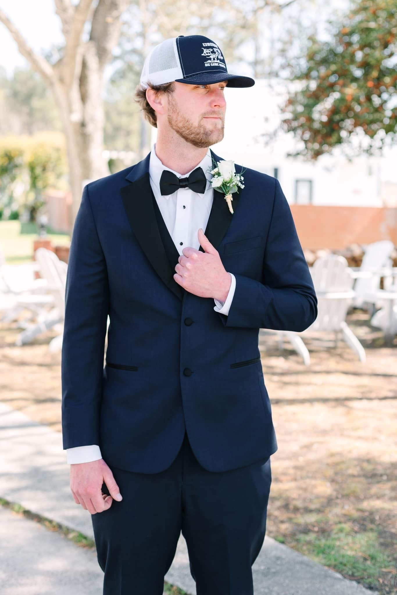 Groom in a navy tuxedo with black bow tie and white boutonniere wearing a trucker cap poses confidently on the grounds of Scarborough House historic estate wedding venue.