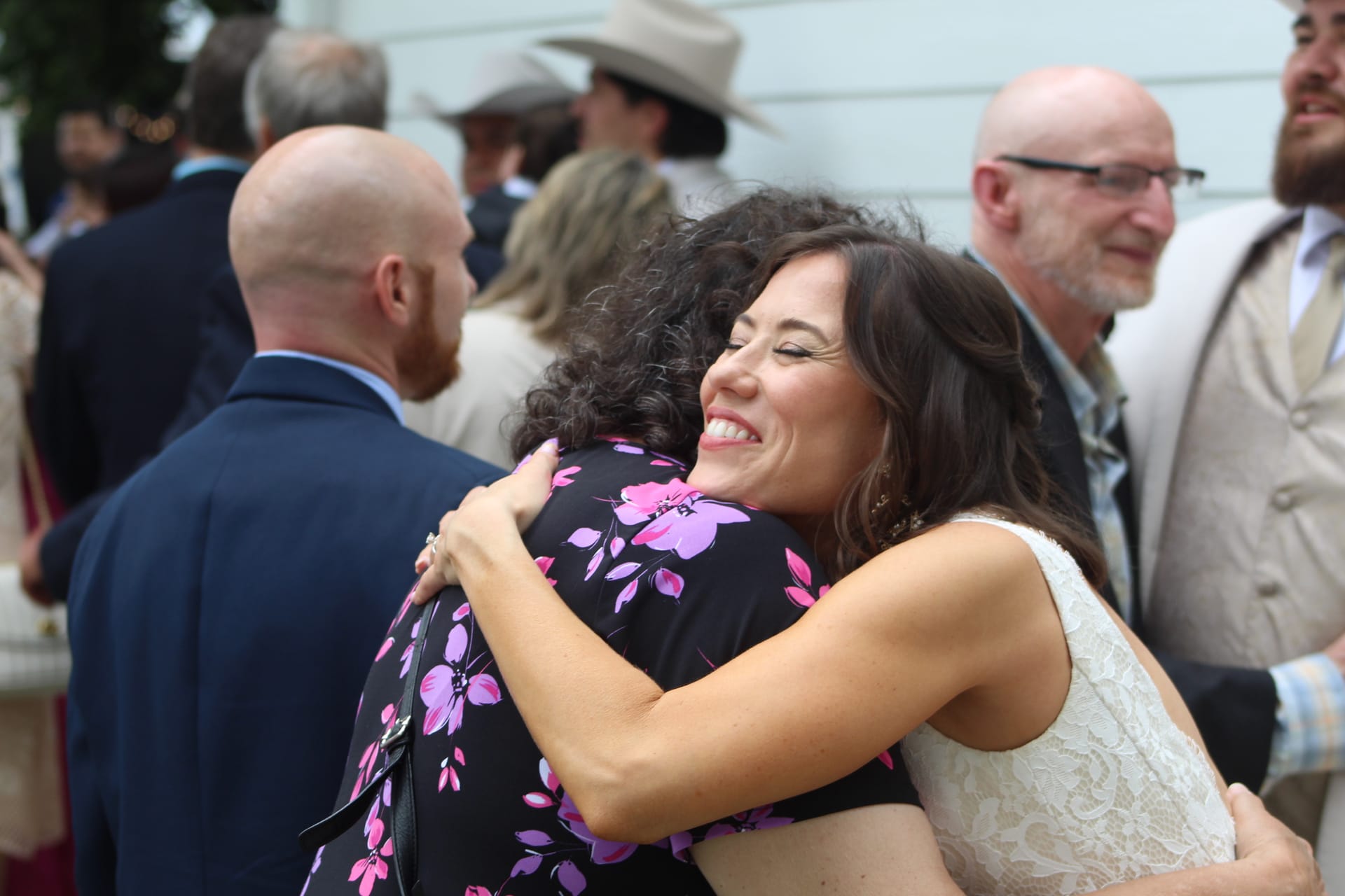 Bride in white lace dress beaming with joy while embracing a guest in a floral dress during an outdoor wedding reception at Scarborough House estate.