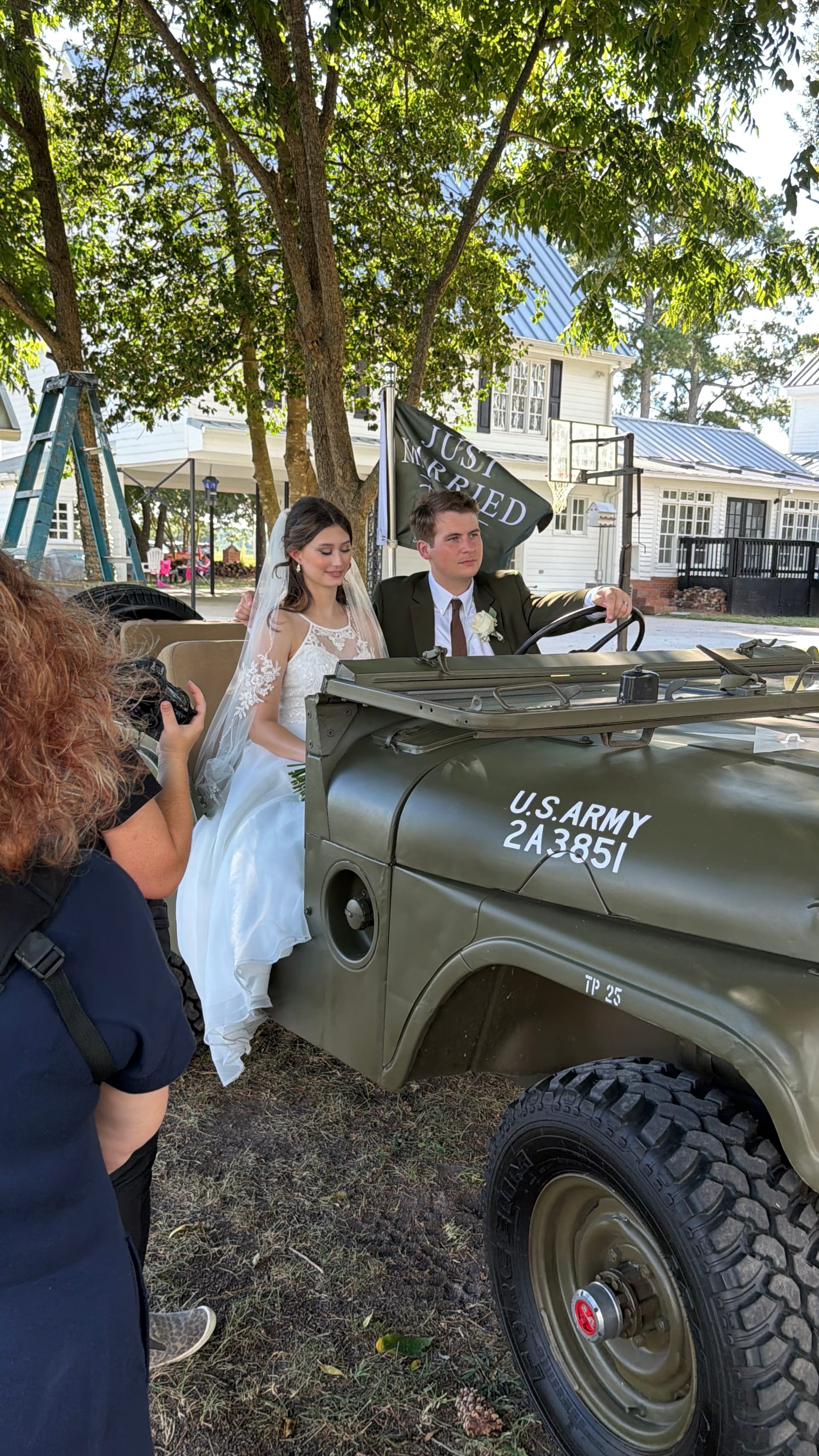 Bride and groom pose in a vintage olive-green U.S. Army Jeep decorated with a Just Married flag beneath oak trees at a historic estate wedding venue.