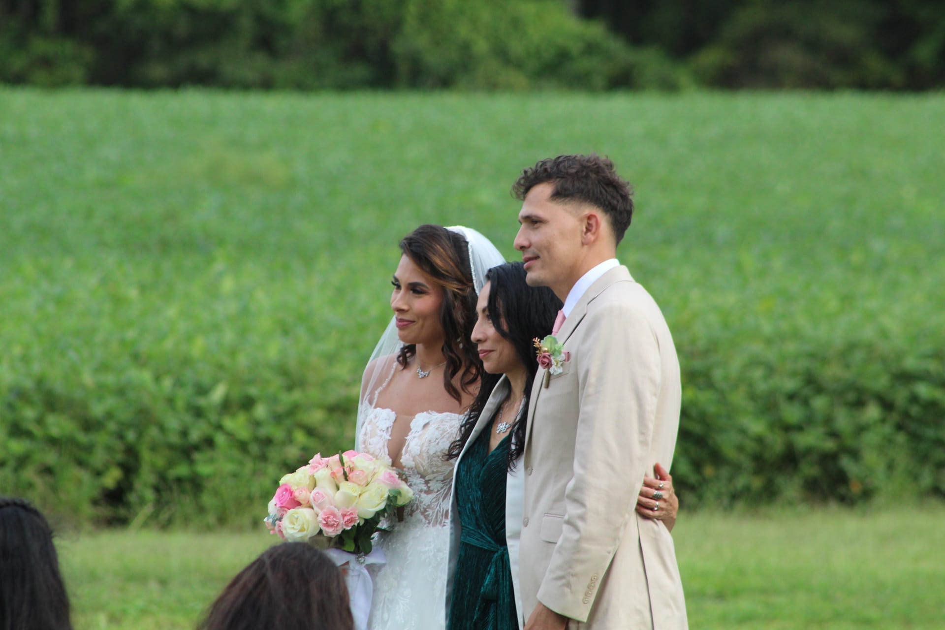 Bride in lace gown holding pink and white bouquet stands with a guest and groom in a tan suit against a sweeping green farmland field at Scarborough House.