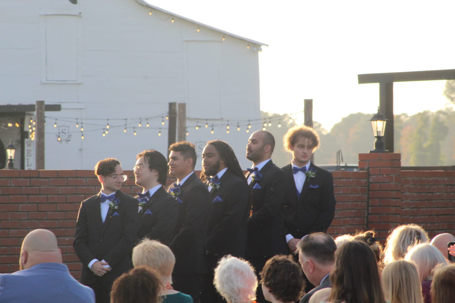 Six groomsmen in black tuxedos with blue bow ties and boutonnieres stand in a row during an outdoor wedding ceremony at Scarborough House with a white barn and string lights in the background.