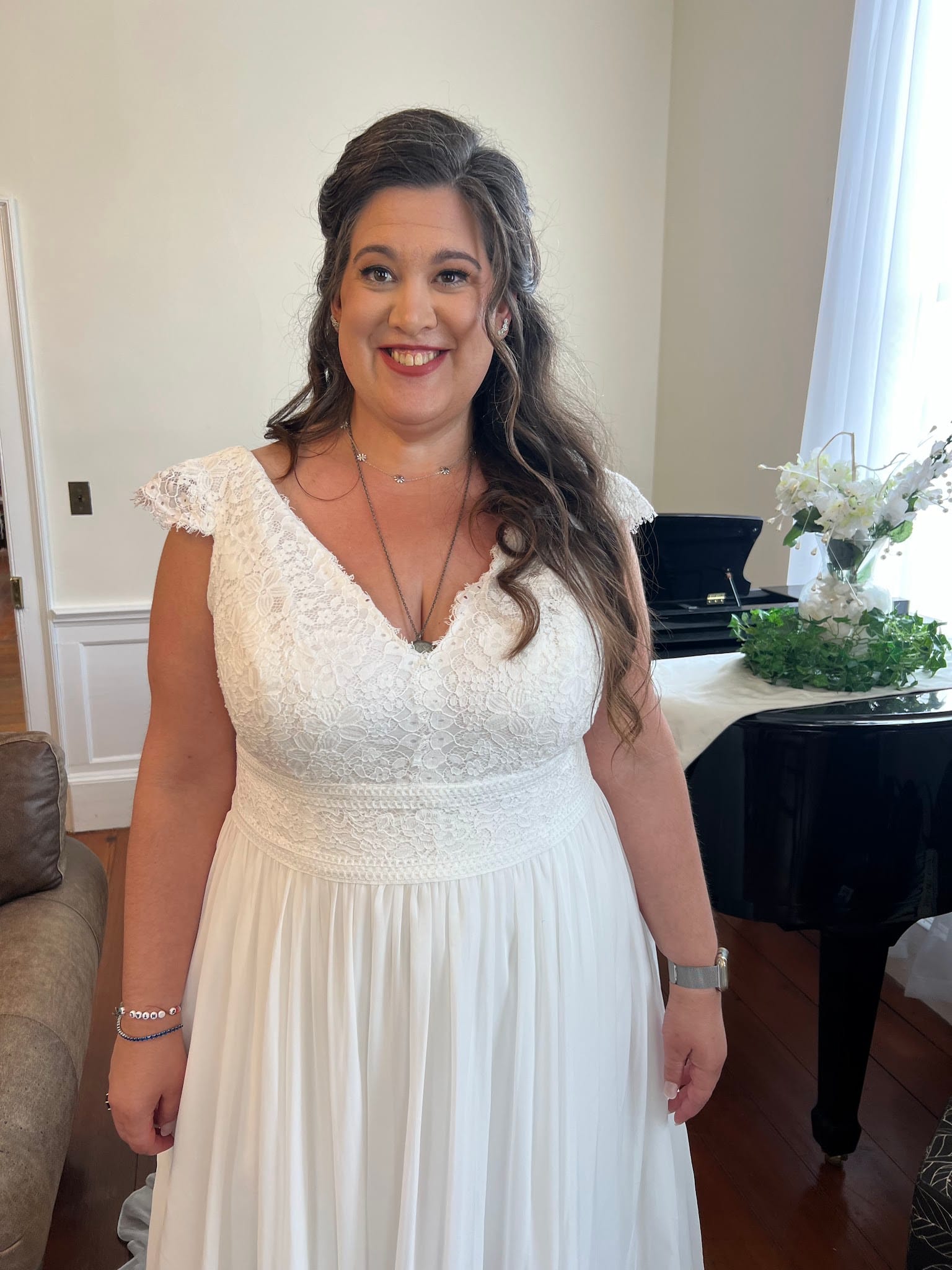 Bride in a white lace cap-sleeve wedding gown smiling warmly inside the historic grand hall at Scarborough House with a black grand piano and white floral arrangement in the background.
