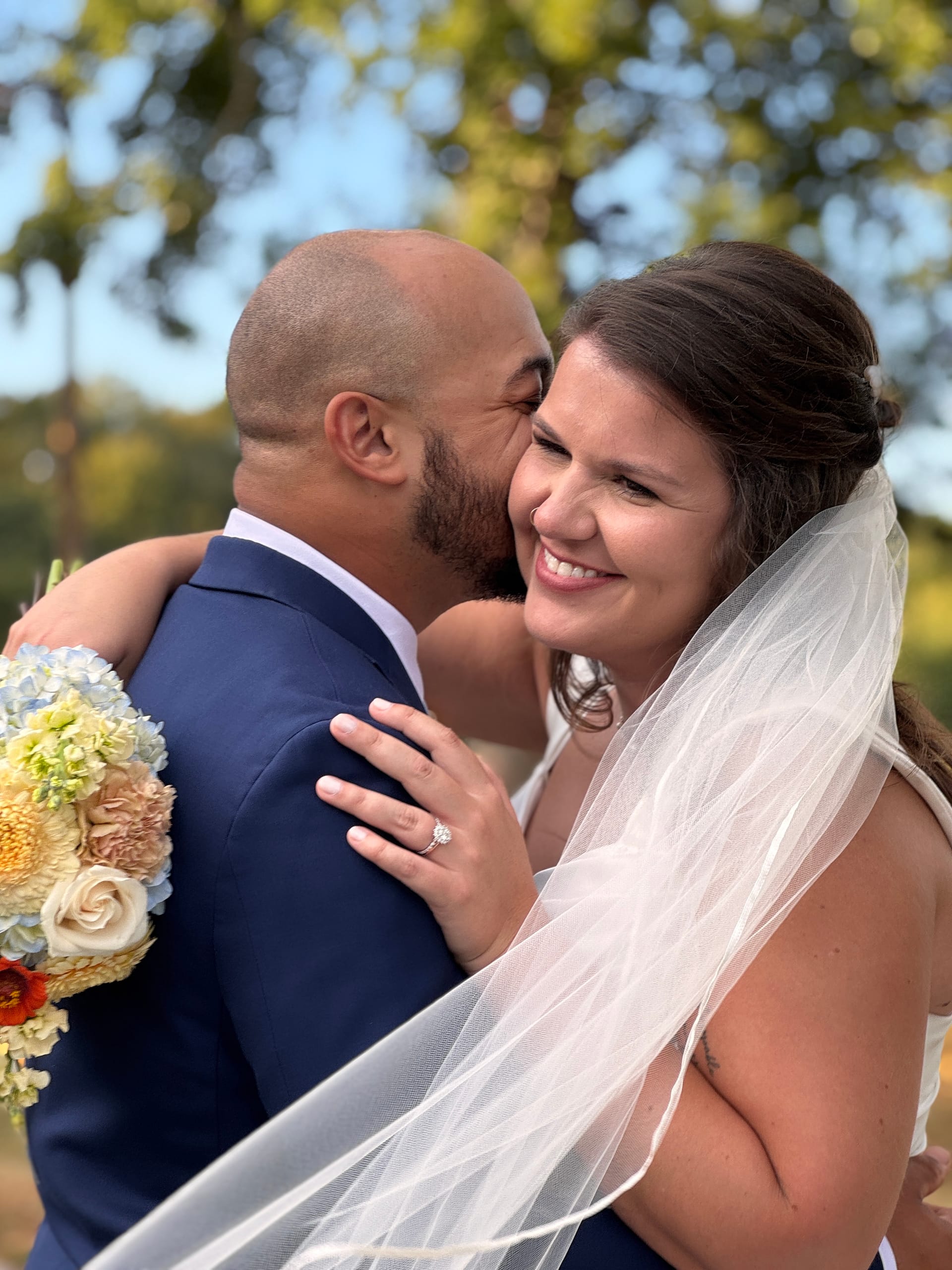 A radiant bride smiles joyfully as her groom kisses her cheek beneath the oak trees at Scarborough House, her veil and colorful bouquet catching the warm natural light.