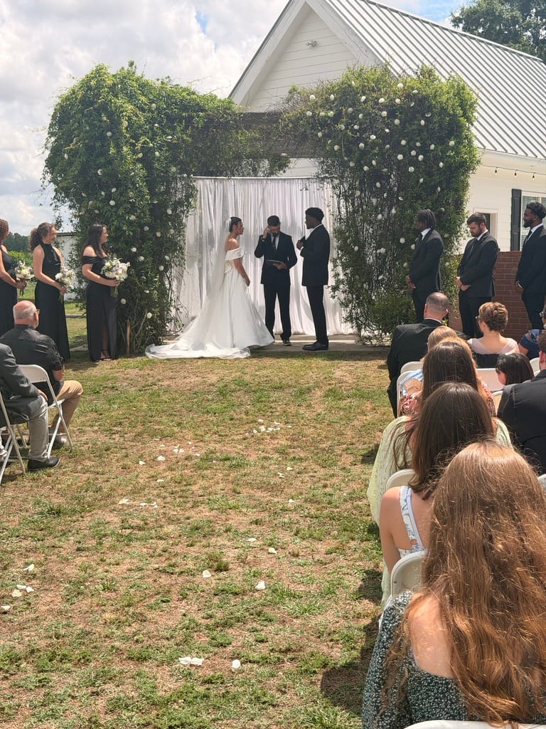 Bride and groom exchange vows beneath a lush greenery and white floral arch with draped fabric backdrop on the ceremony lawn at Scarborough House, surrounded by guests and wedding party.