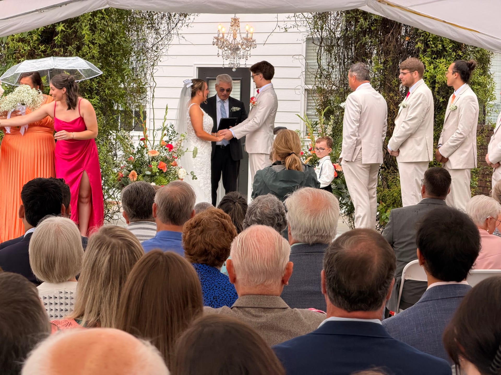 A bride and groom exchange vows beneath an ivy-covered arch with a crystal chandelier at Scarborough House, surrounded by their wedding party in blush and coral attire and seated guests.
