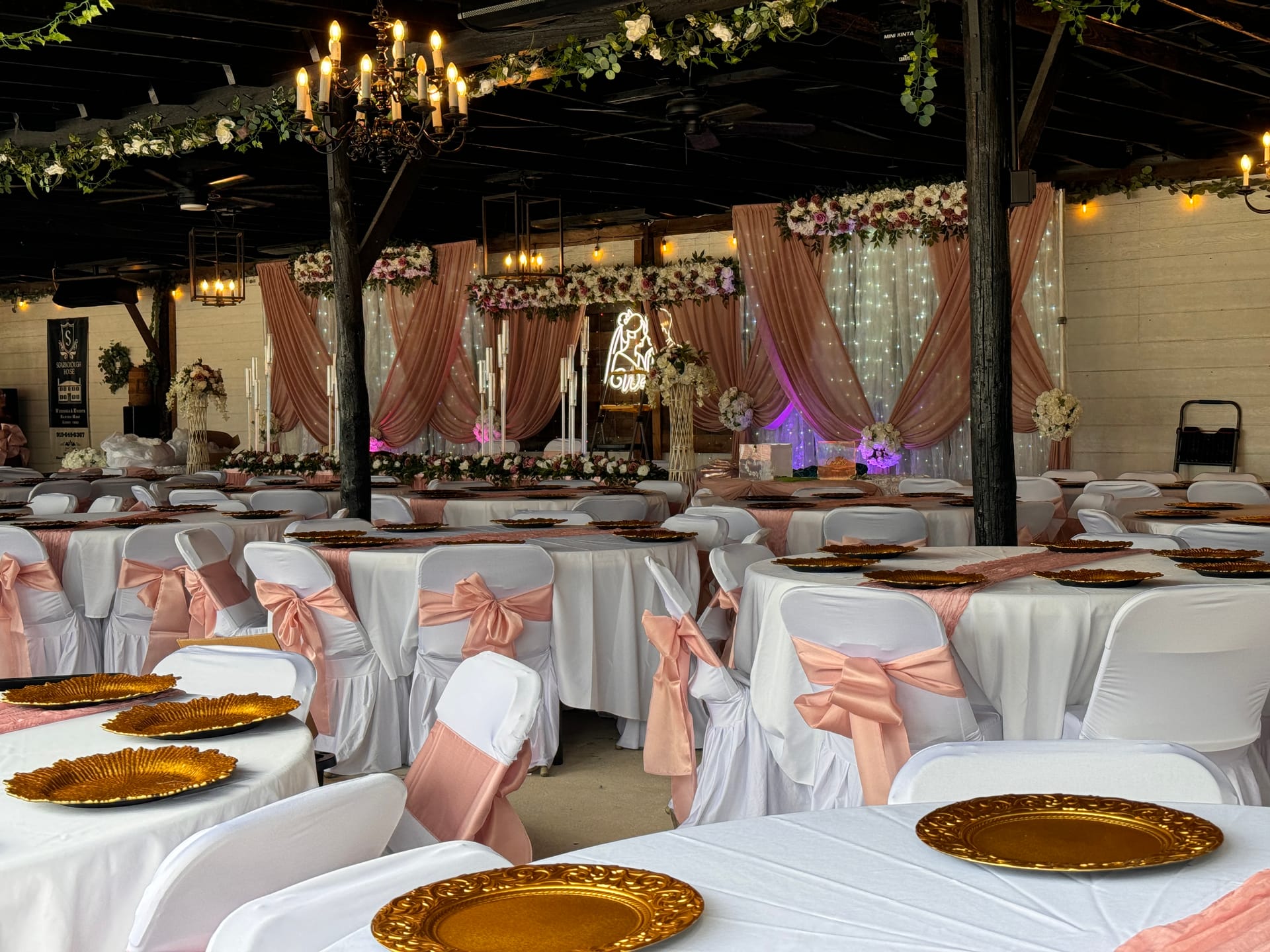 Elegantly decorated wedding reception hall at Scarborough House featuring blush pink chair sashes, gold charger plates, floral arrangements, draped fabric backdrop, and warm chandelier lighting.