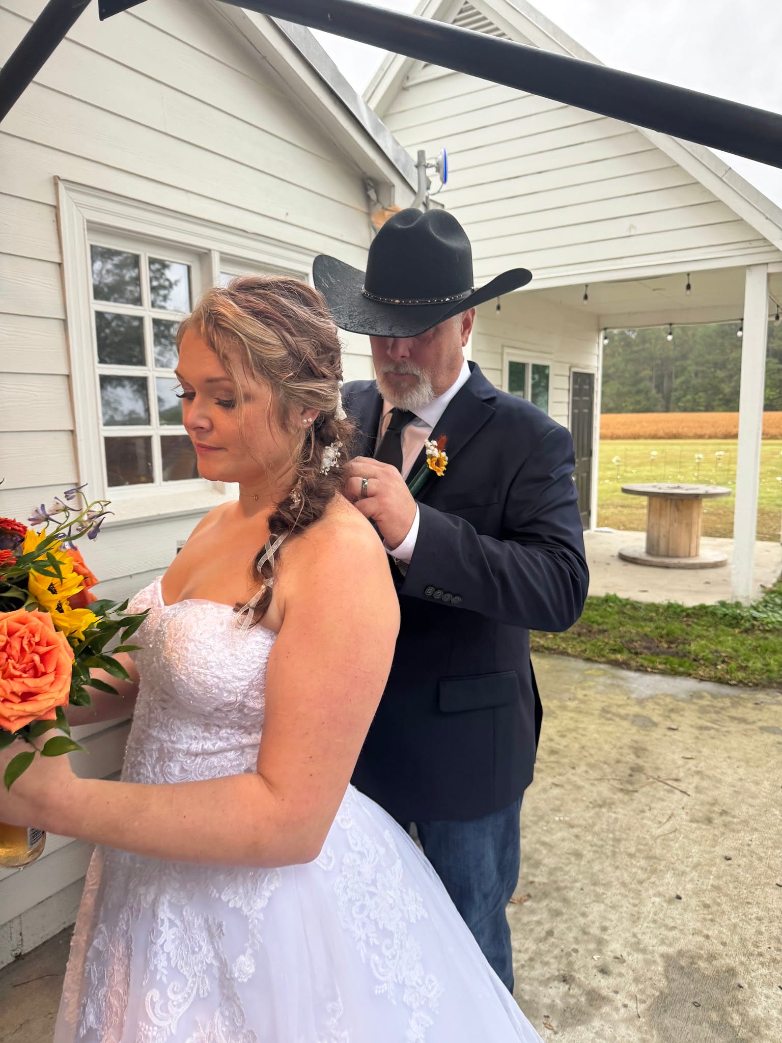 Groom in black cowboy hat and navy blazer adjusts his boutonniere while standing behind his bride in a white lace gown holding a colorful bouquet outside a white historic estate building.
