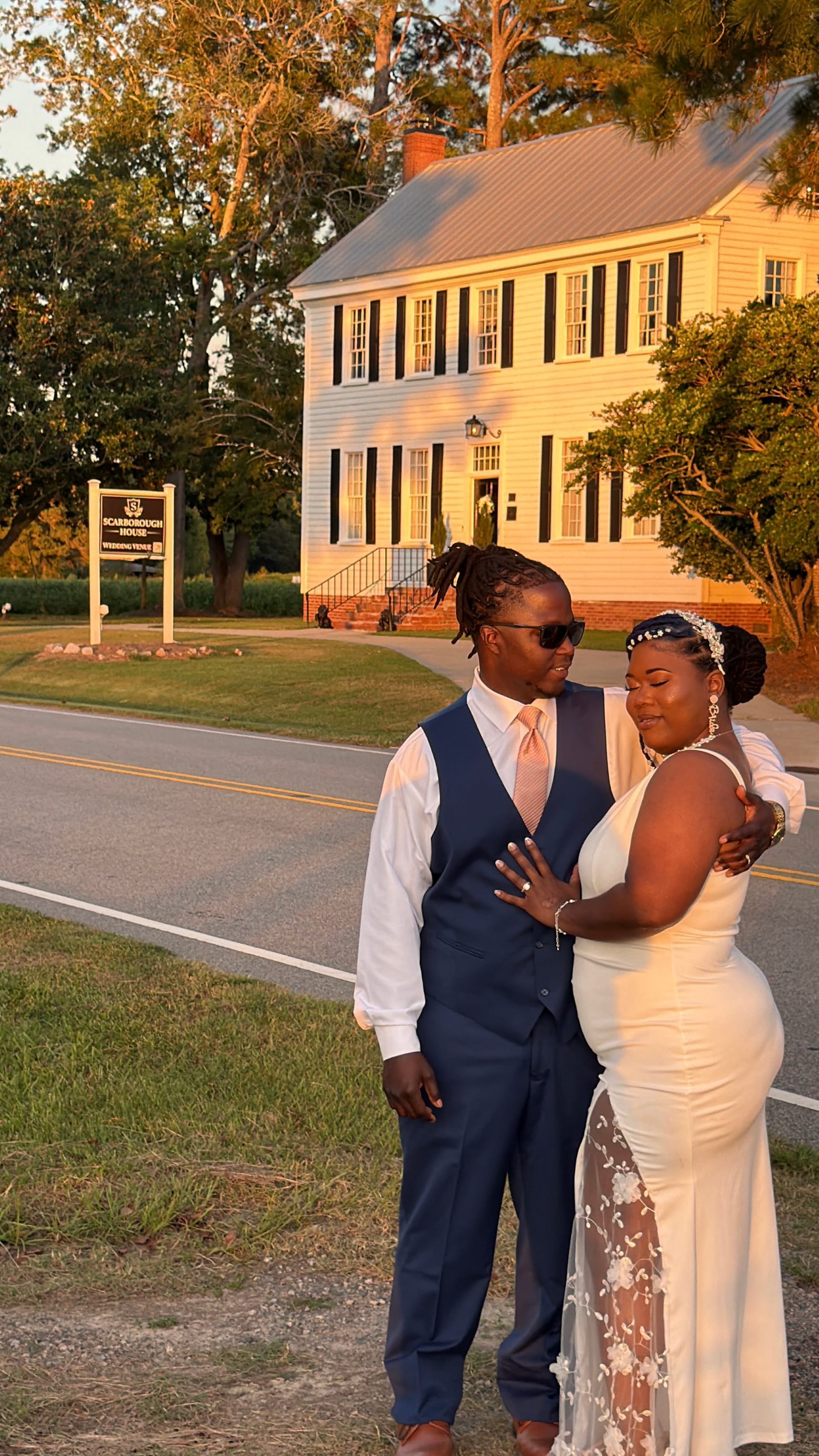 Bride in white gown and groom in navy vest share a tender embrace on the road in front of the historic 1821 Scarborough House manor during golden hour.