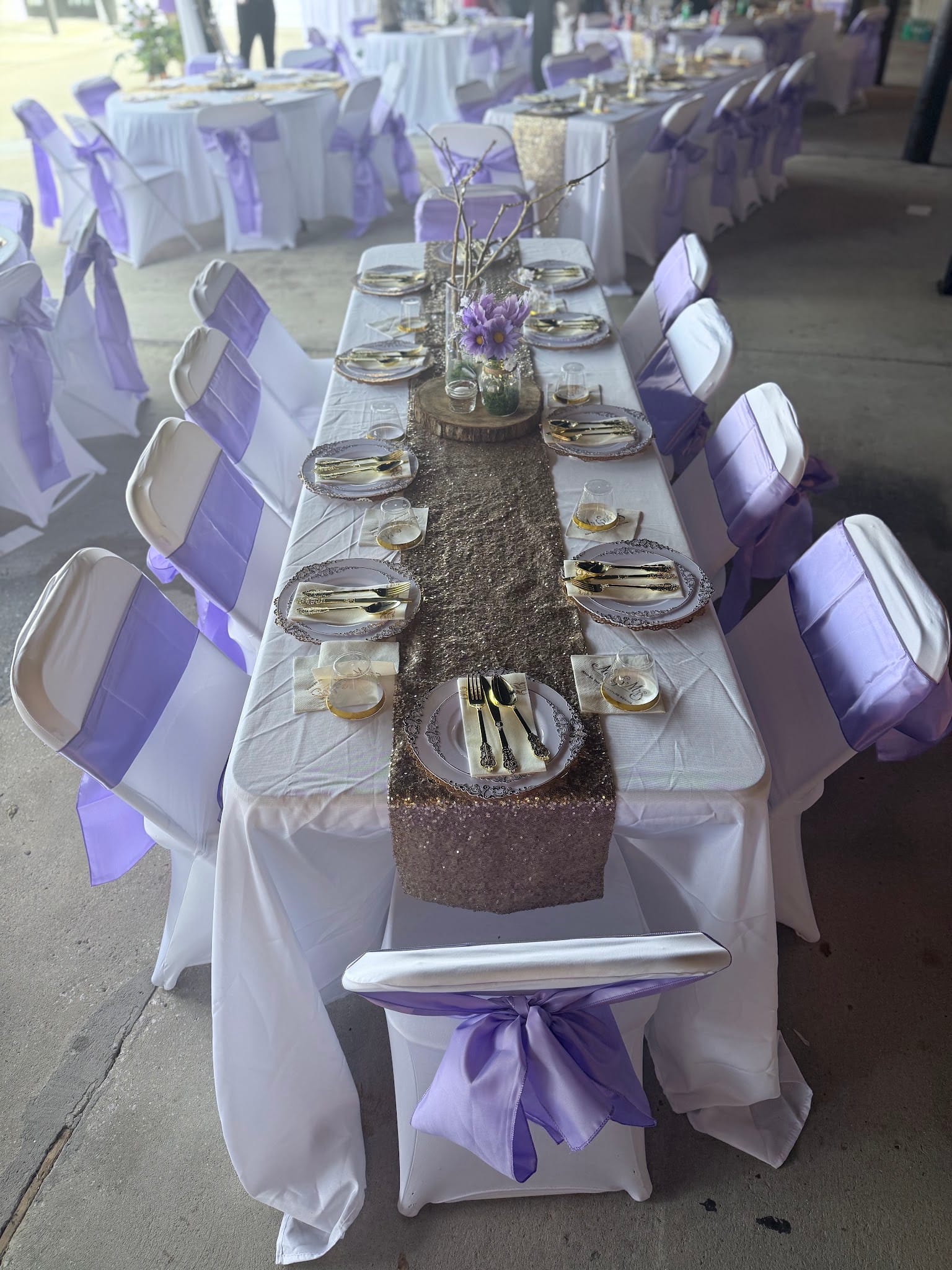 Elegantly set banquet table with a gold sequin runner, lavender chair sashes, ornate charger plates, gold cutlery, and a floral centerpiece at a wedding reception.