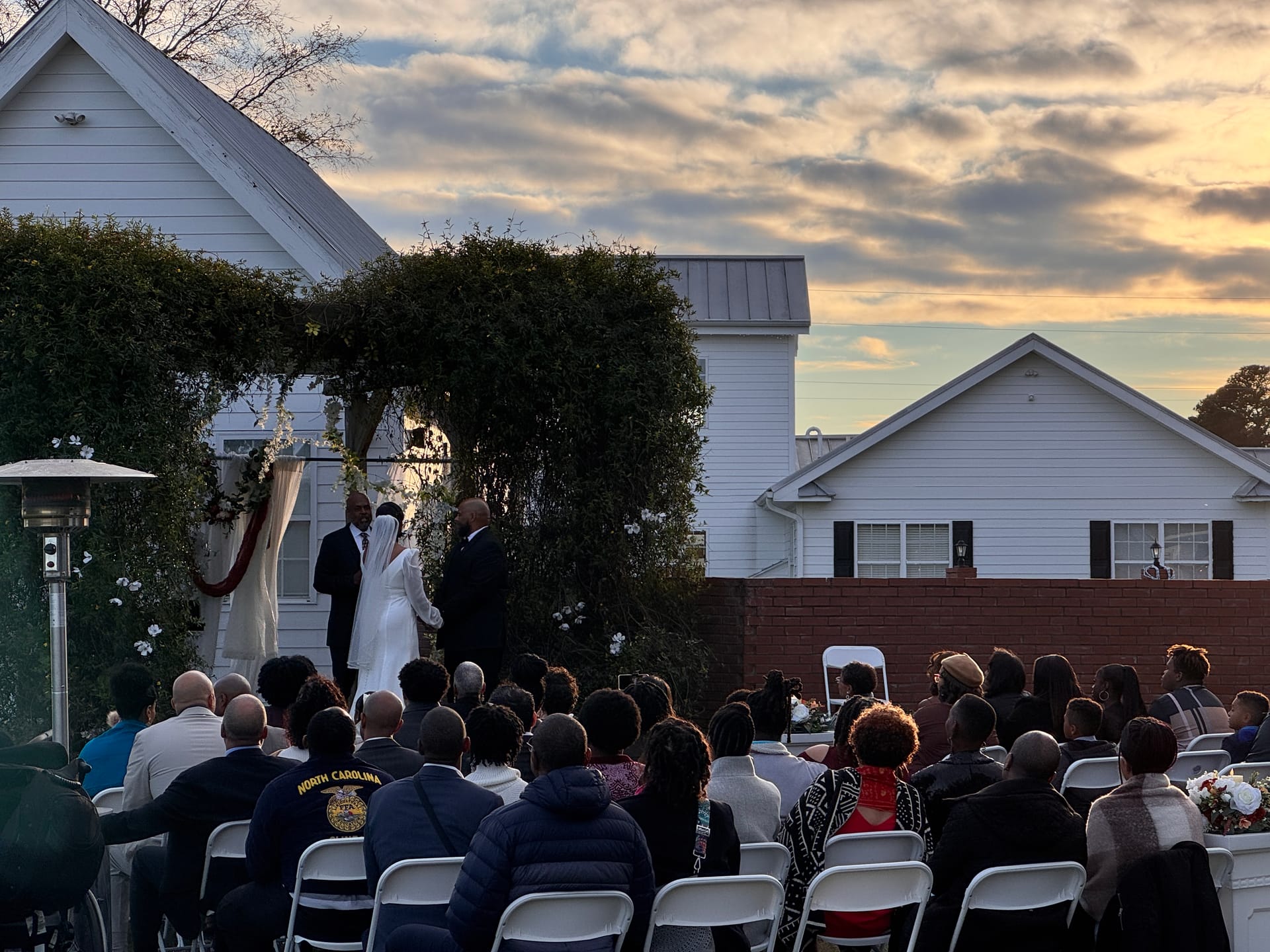 Guests seated in white folding chairs watch a couple exchange vows beneath an ivy-covered archway at dusk, with the historic white manor house and a dramatic golden sunset sky in the background.
