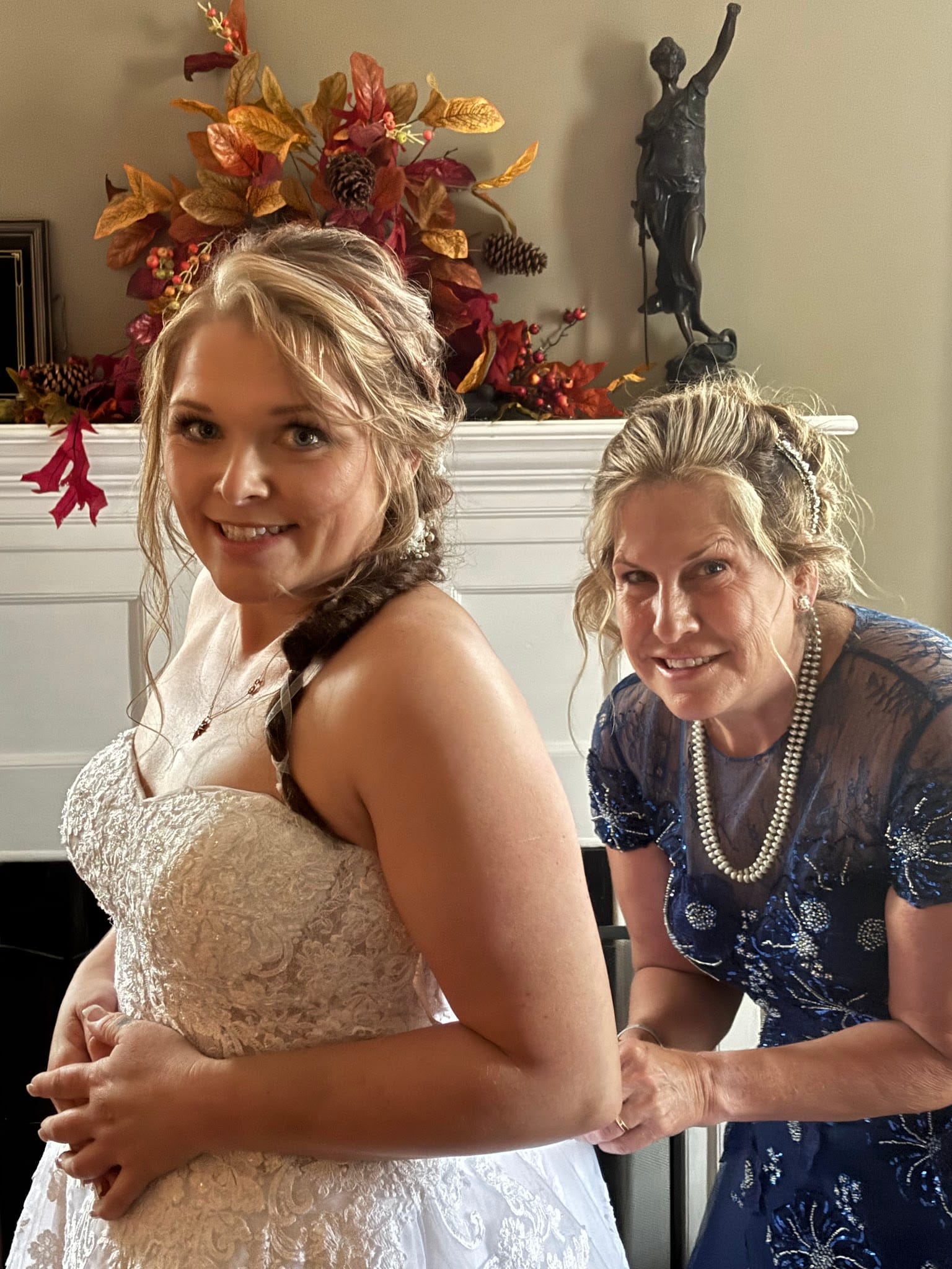 A smiling bride in a white lace gown looks over her shoulder as an older woman in a blue beaded dress fastens the back of the dress in front of a fall-decorated fireplace mantel.