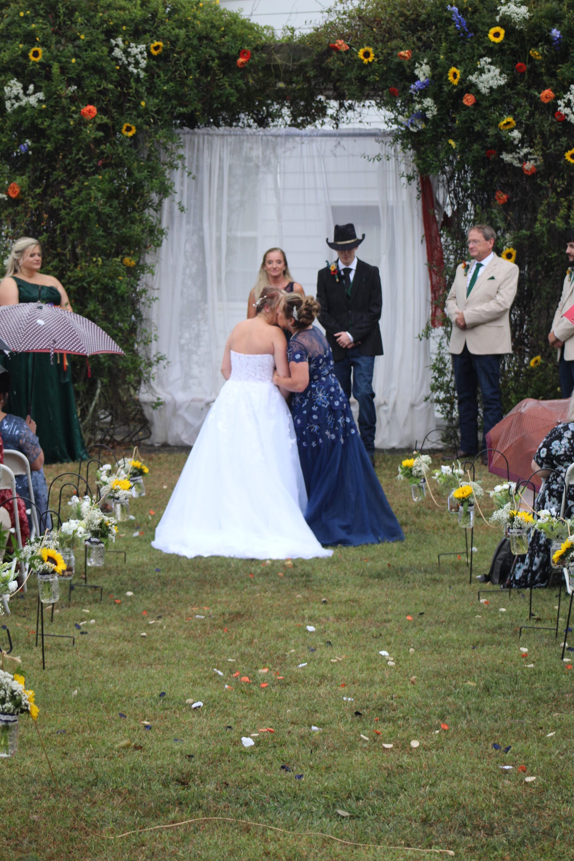 Bride sharing an emotional embrace at the altar during an outdoor garden wedding ceremony framed by a floral arch decorated with sunflowers at a historic estate venue.