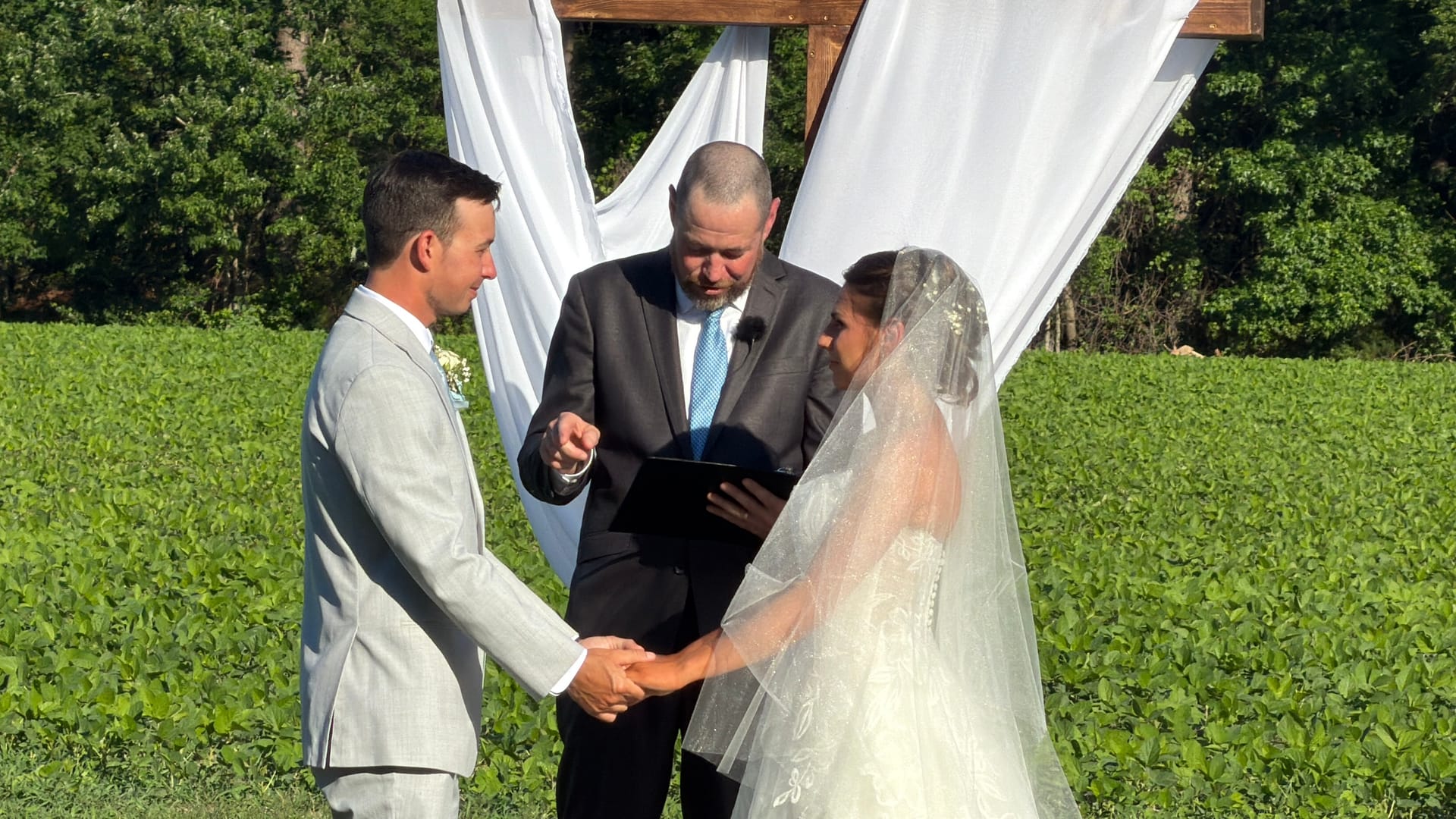 Bride and groom hold hands during outdoor vows beneath a rustic wooden arch draped in white fabric with a lush green field in the background at Scarborough House.